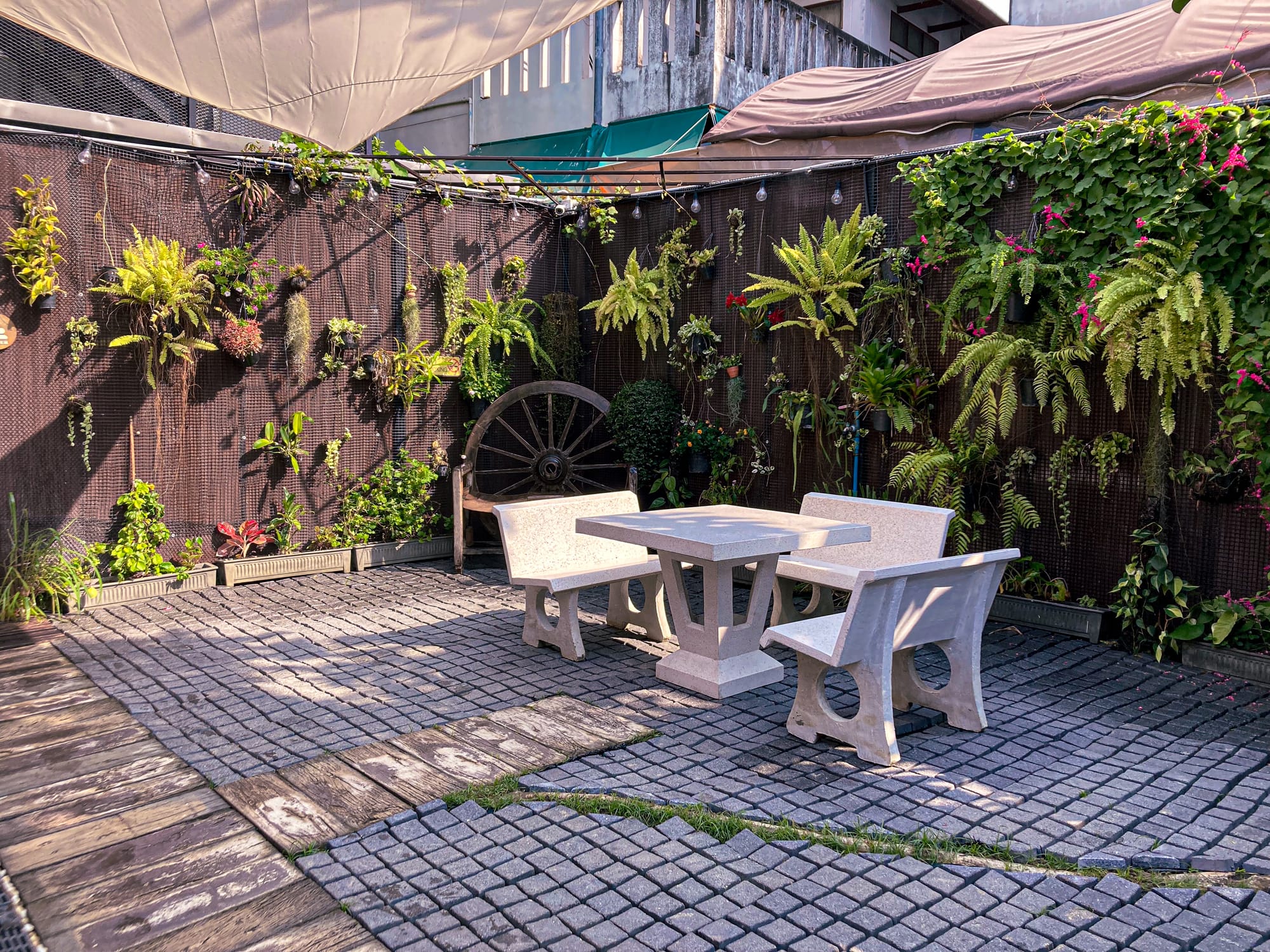 Outdoor seating area at Old City Spa in Chiang Mai featuring stone benches and a table surrounded by lush greenery, hanging plants, and a rustic wooden wheel accent against a garden wall