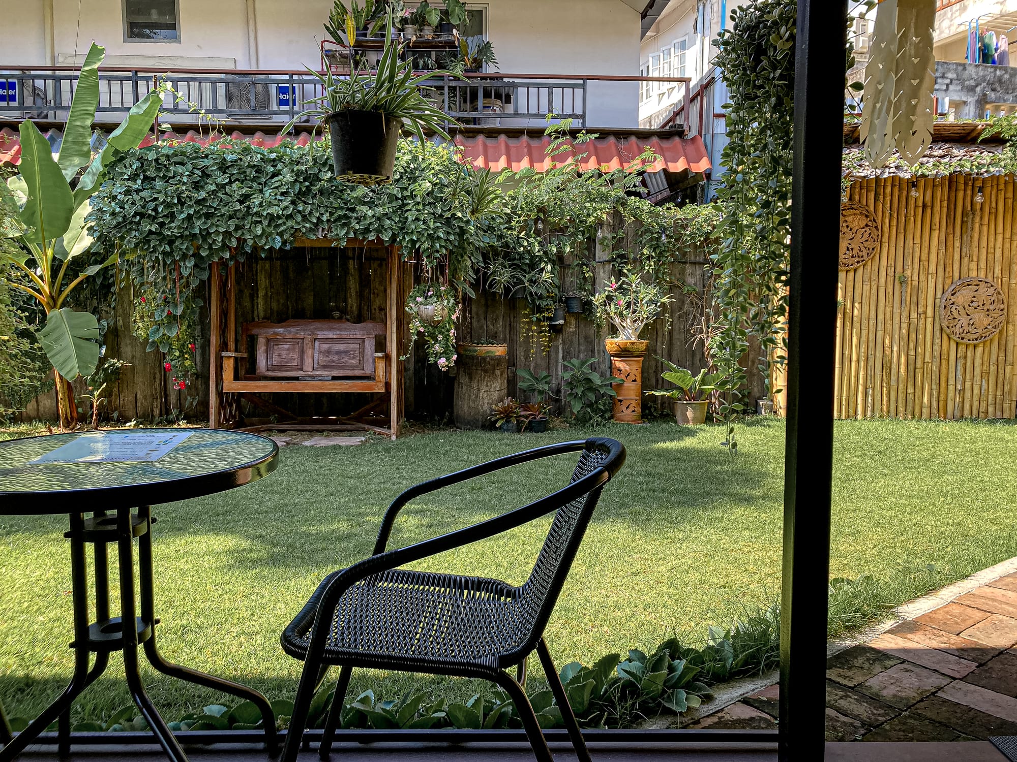 Outdoor garden seating area at Old City Spa in Chiang Mai, featuring a black metal chair and table overlooking a lush green lawn, potted plants, and a wooden swing bench framed by vines
