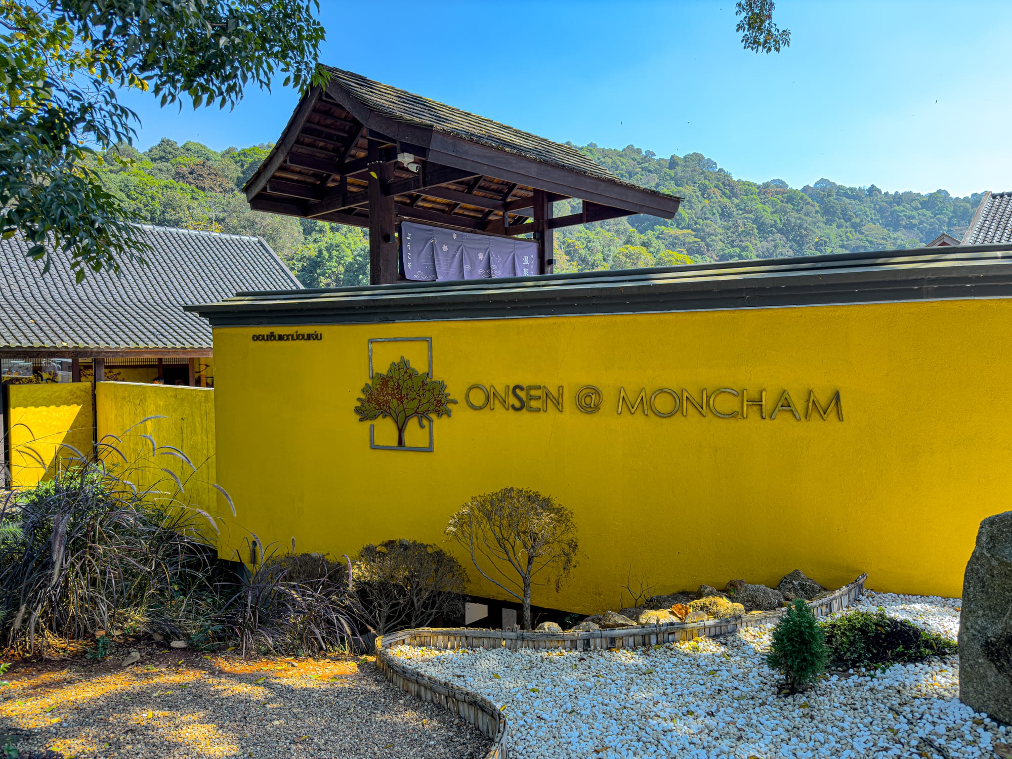 Yellow exterior wall with Onsen at Moncham sign, traditional roof and garden landscaping in Mae Rim, Thailand