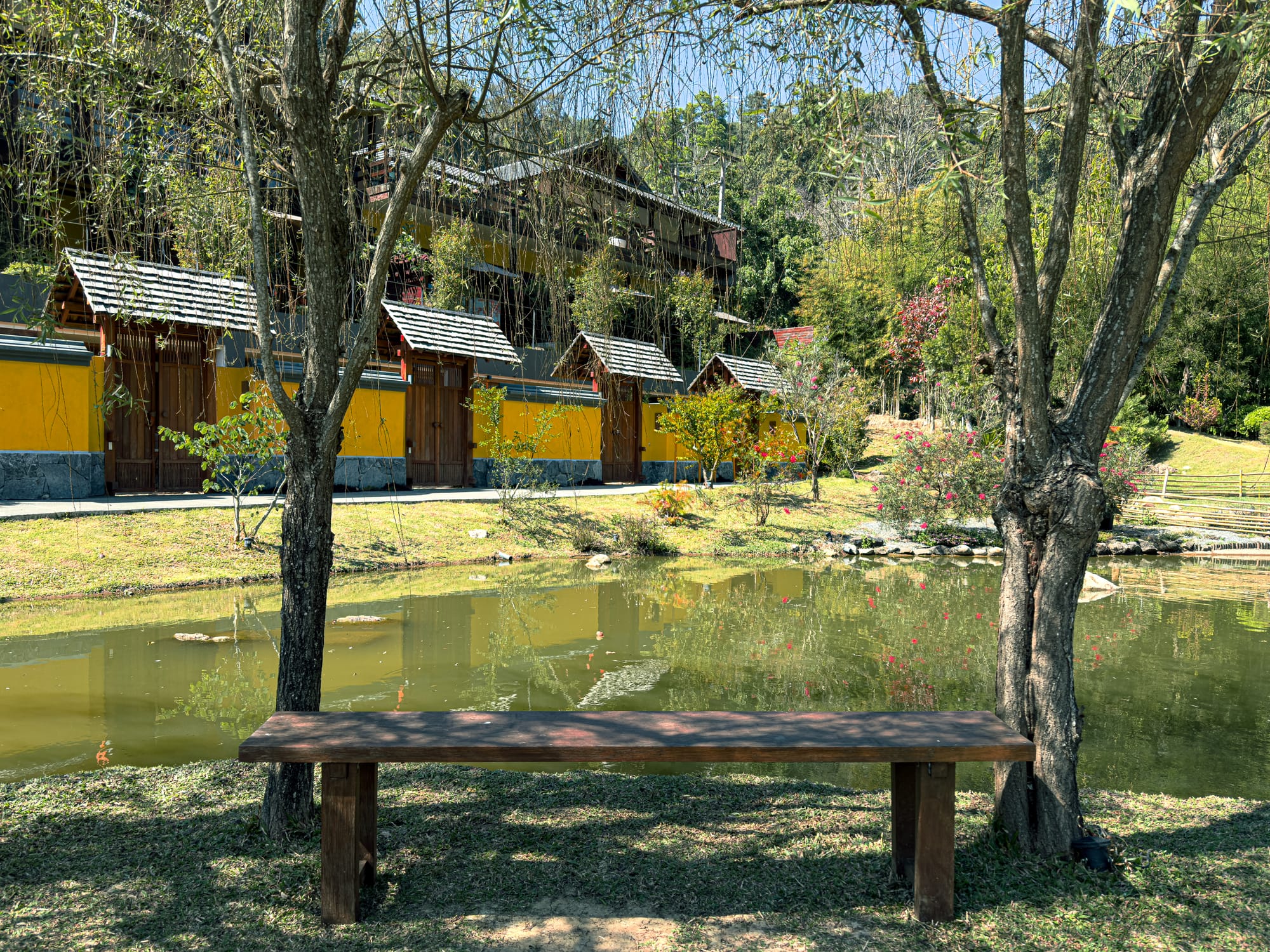 A tranquil wooden bench beneath two trees overlooking a pond, with yellow-walled onsen buildings and lush greenery in the background at Onsen at Moncham
