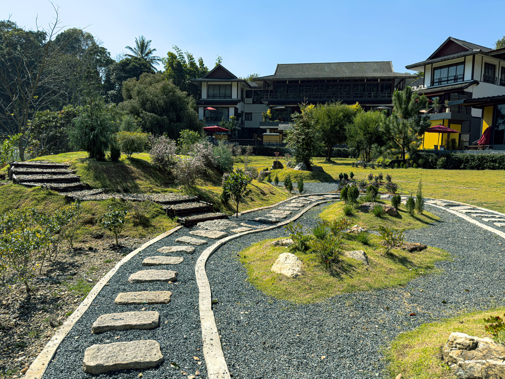 Stone pathways winding through landscaped gardens with shrubs and trees, leading toward traditional-style buildings at Onsen at Moncham in Mae Rim, Thailand