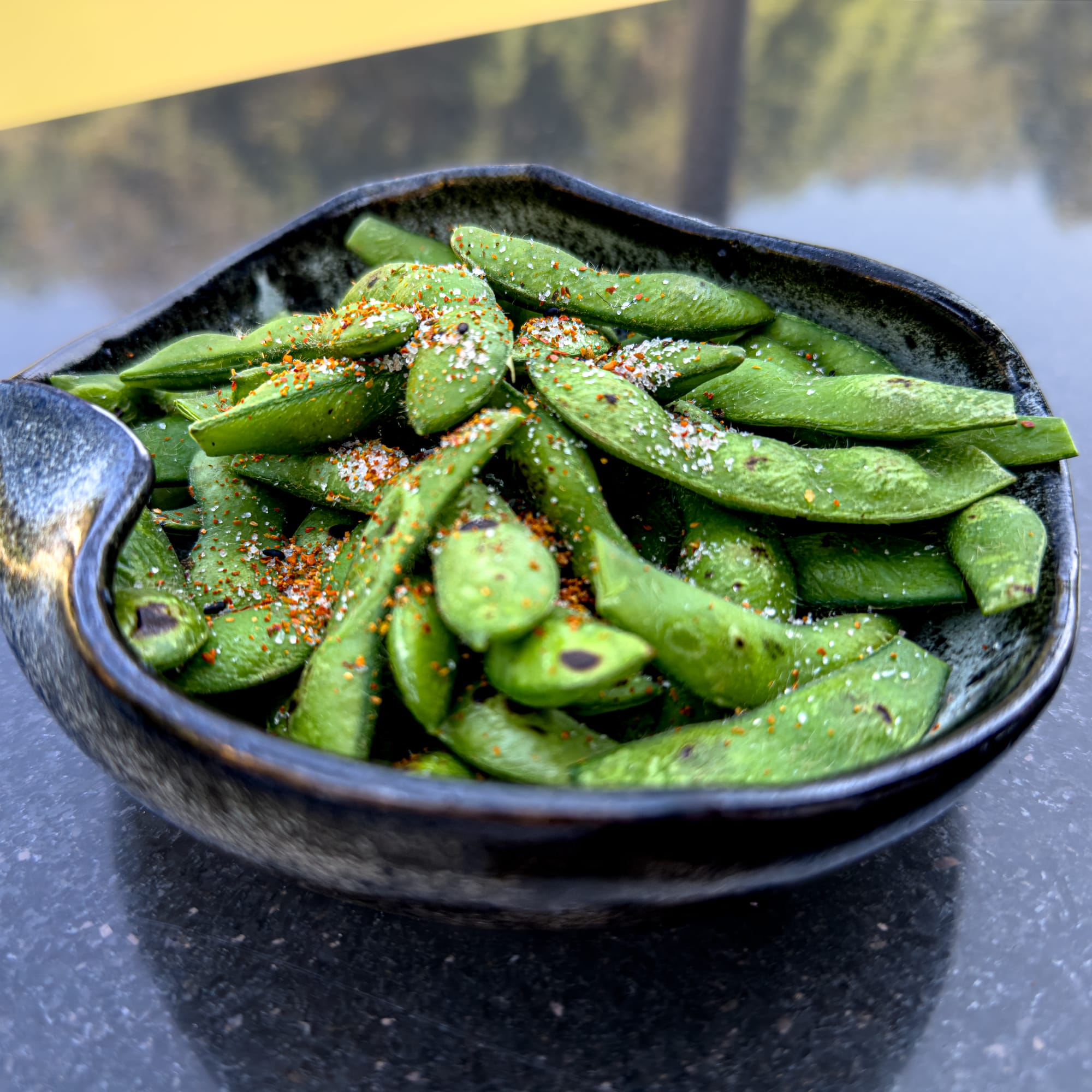 A close-up of a black ceramic bowl filled with edamame pods sprinkled with salt and chili seasoning, placed on a reflective dark surface