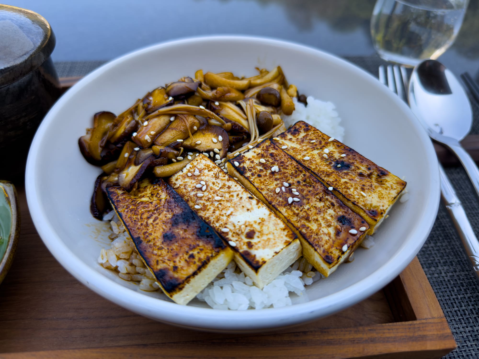 A close-up of a Japanese dish featuring grilled tofu slices, sautéed mushrooms, sesame seeds, and rice in a white bowl, served on a wooden tray with cutlery in the background