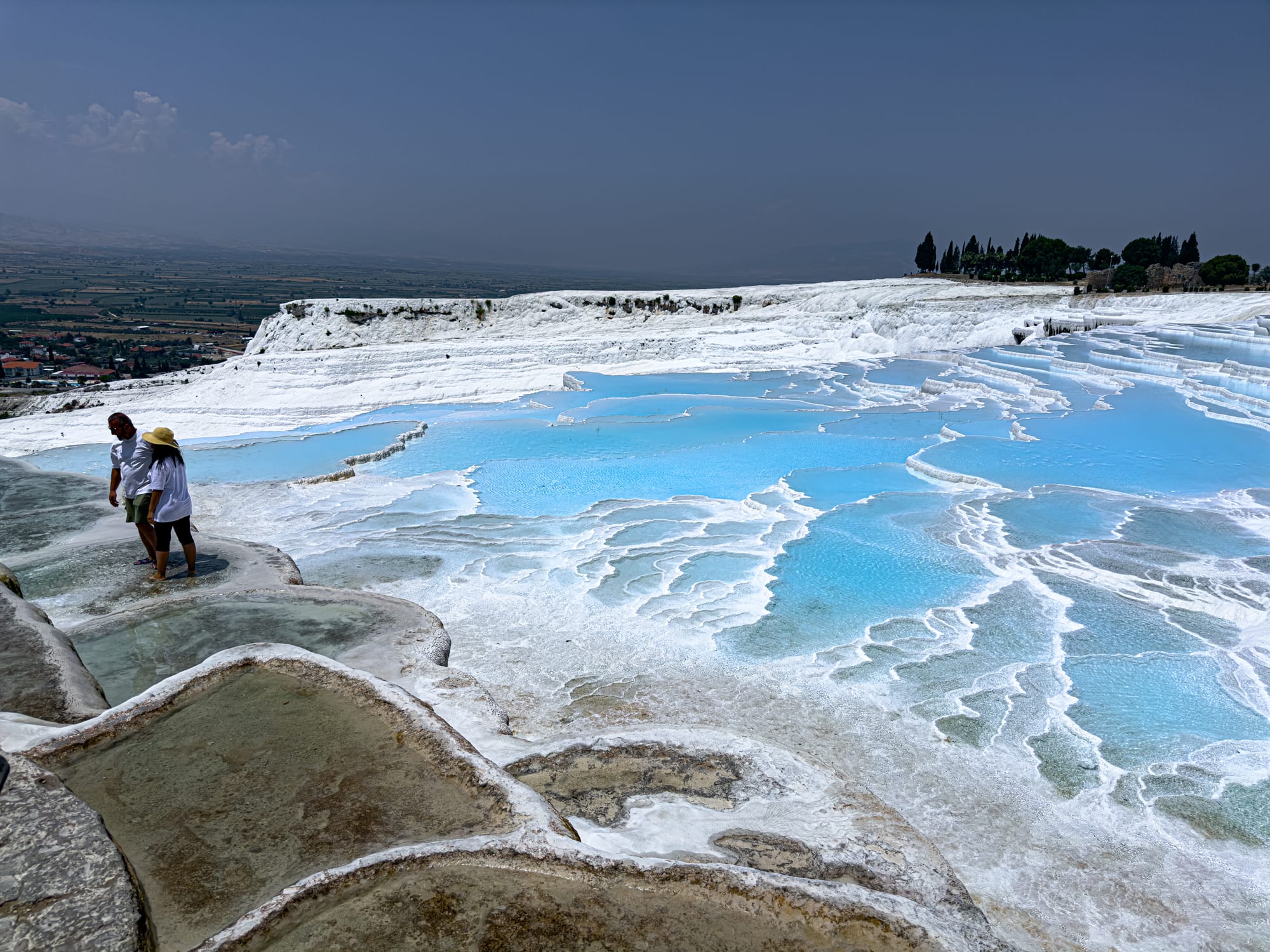 Two people walking across Pamukkale’s white travertine terraces with turquoise thermal pools under a clear sky