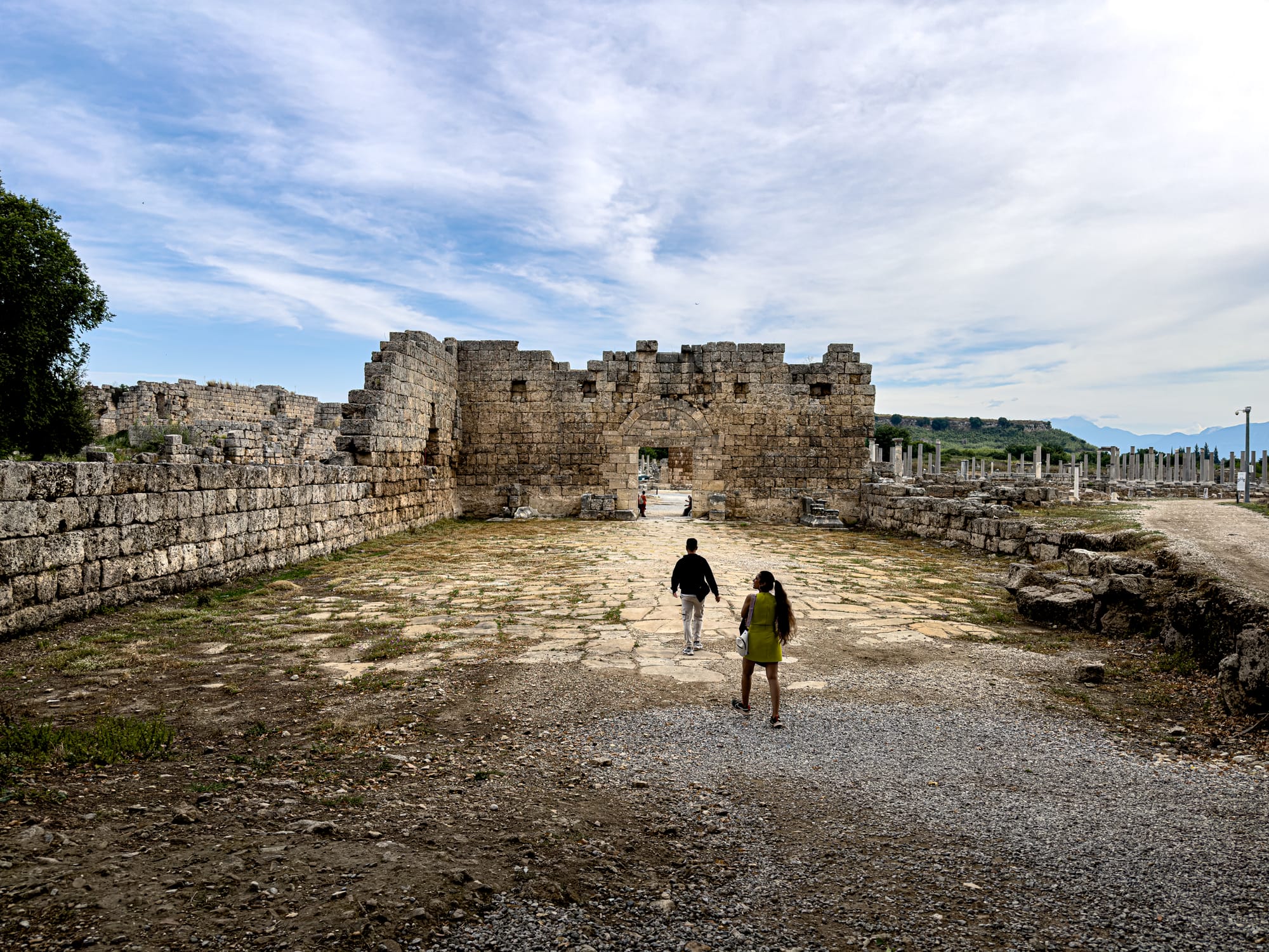 Visitors walking toward the stone entrance of the gate at Perge with column-lined streets in the background near Antalya, Turkey
