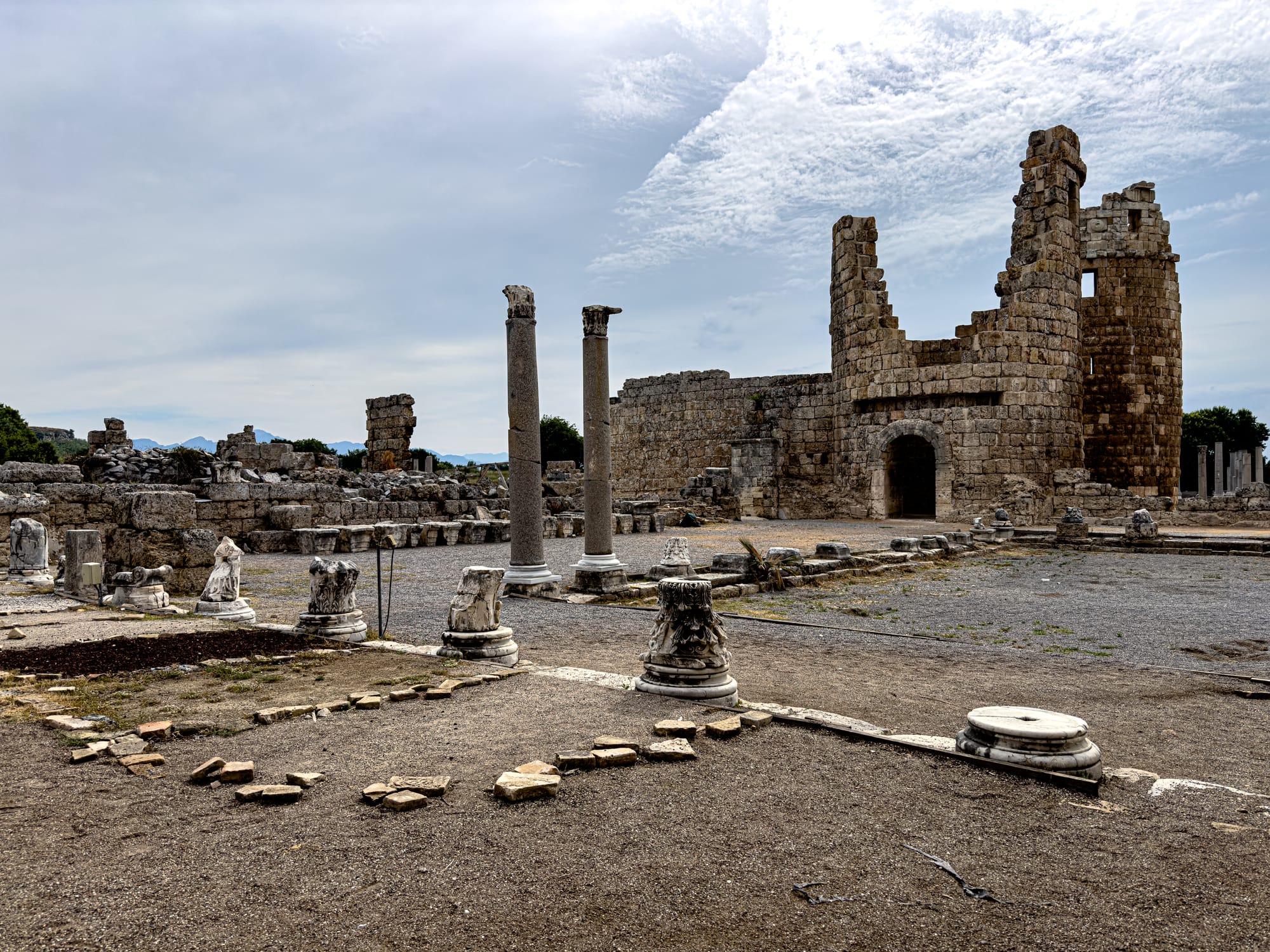 Wide view of the Hellenistic Gate and scattered column bases at Perge with ruins and stone walls under a partly cloudy sky near Antalya, Turkey