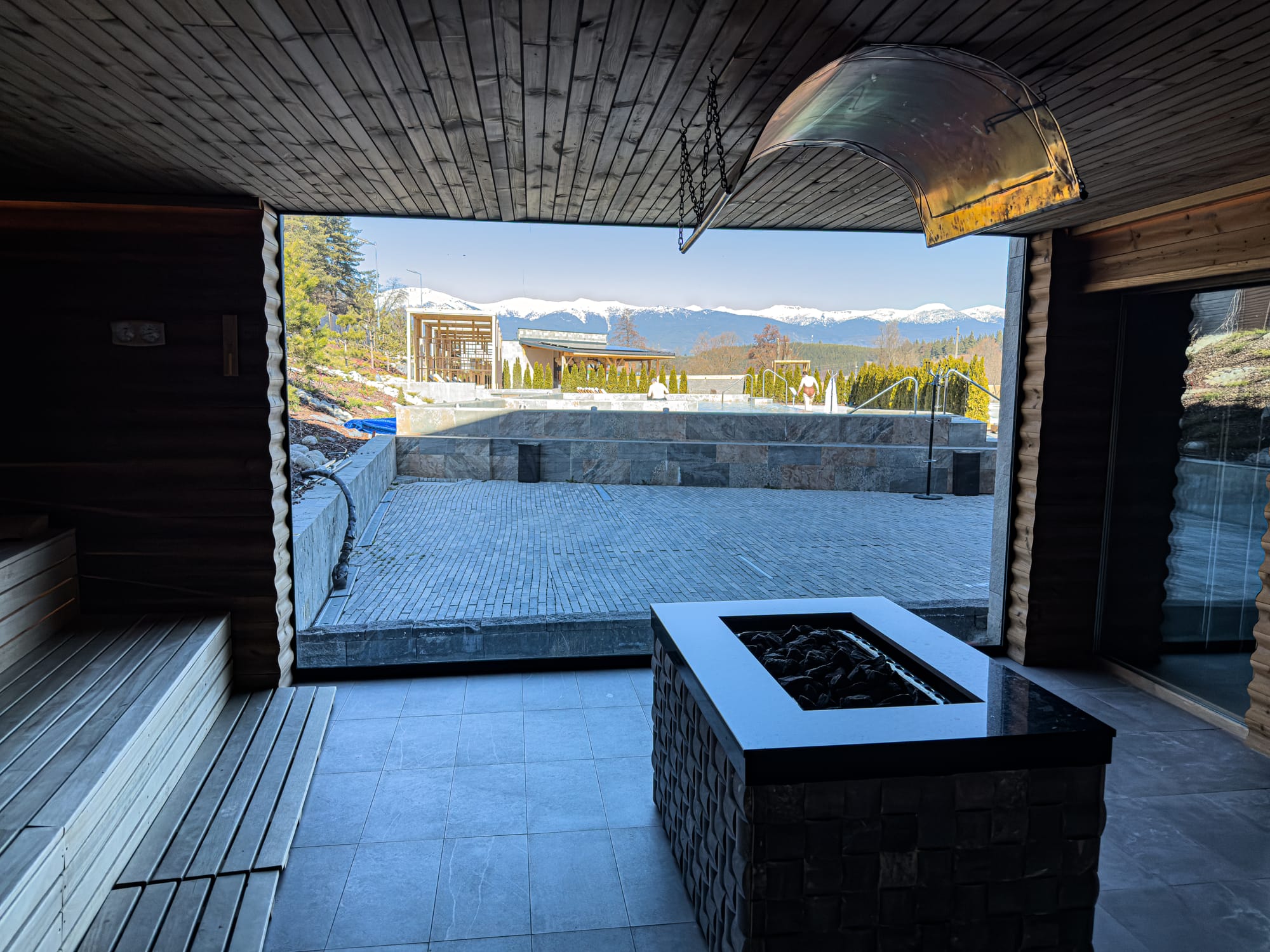 Inside a wooden sauna at Pulse Thermal Spa in Banya, Bulgaria, with a heater in the foreground and large window framing outdoor pools and the snowy Pirin mountain range in the distance