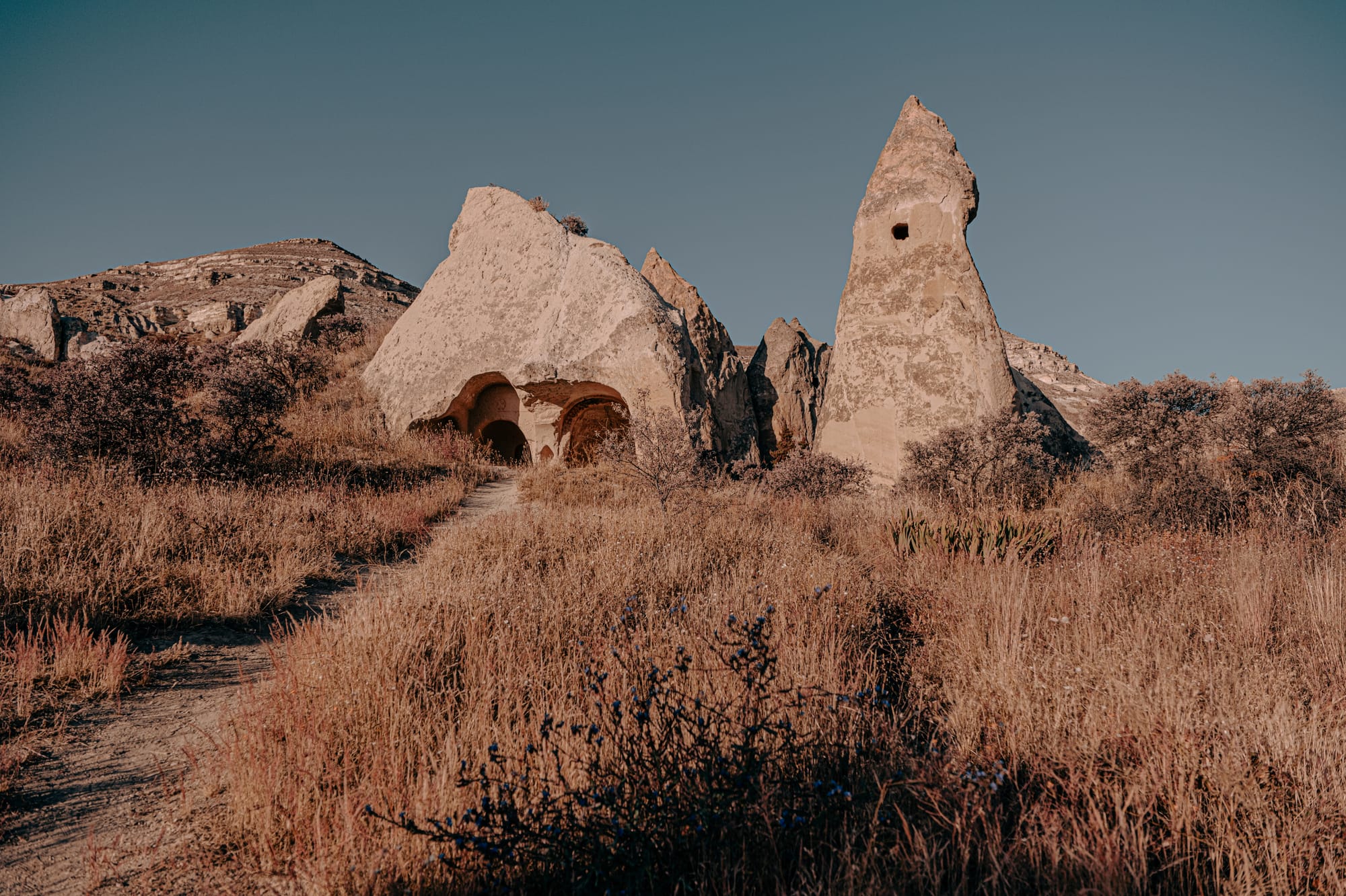A view of two prominent rock formations in Cappadocia, one with a pointed peak and small window-like opening, the other containing two arched cave entrances, surrounded by dry grass and shrubs under a clear blue sky