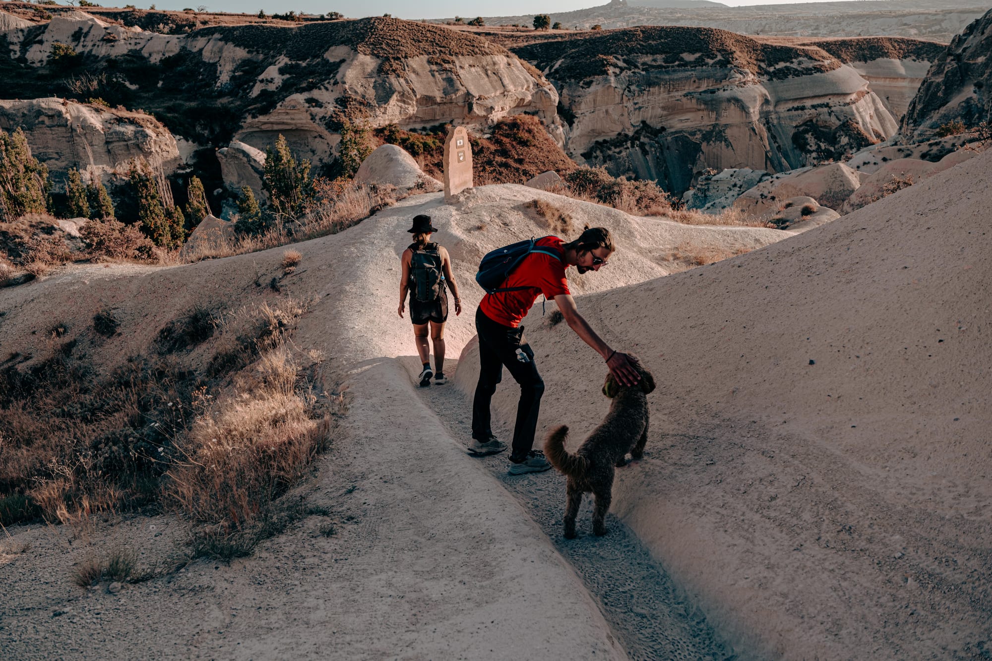 A man in a red shirt and sunglasses bends down to pet a brown curly-haired dog named Oscar while walking along a narrow dirt trail in Cappadocia, with another hiker ahead and dramatic canyon cliffs in the background