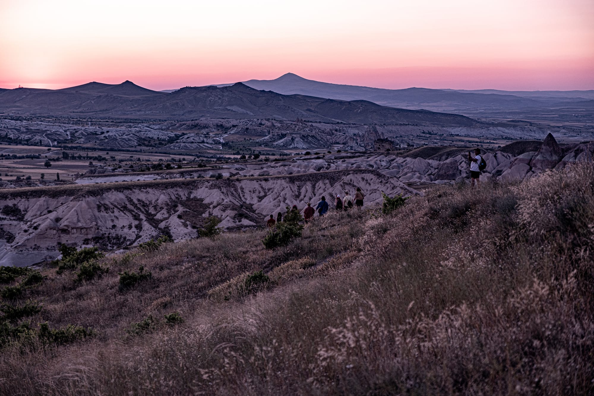 A group of hikers walking along a grassy ridge at sunset in Cappadocia, with the Red and Rose Valleys stretching out in the distance, their rock formations tinted purple and pink under the fading light, and a lone figure pausing to take a photo of the landscape