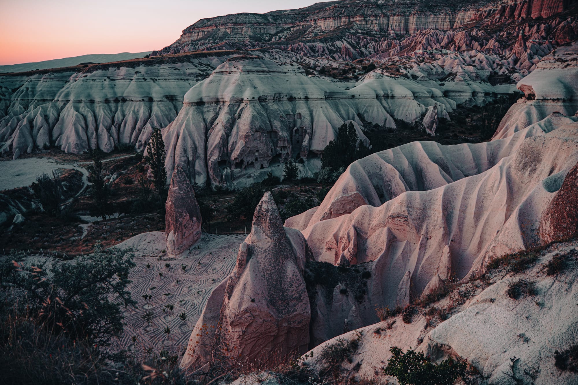 Panoramic view of Cappadocia’s Red and Rose Valleys at sunset, showing dramatic ridges, soft pink and grey rock formations, and pointed fairy chimneys, with carved openings visible in the cliff faces and agricultural patterns in the valley floor below