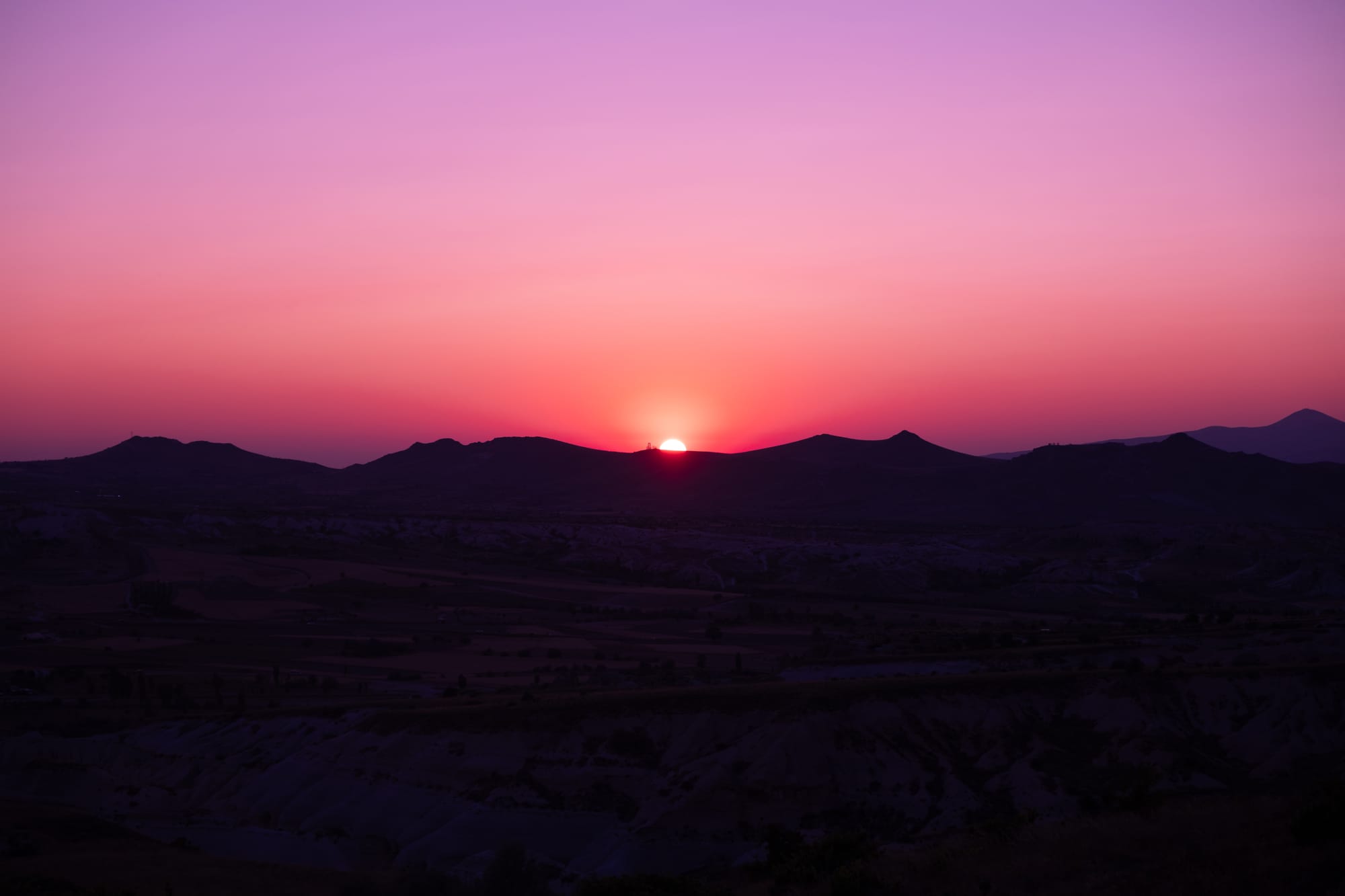 The sun setting behind Cappadocia’s distant hills, casting the sky in rich gradients of pink, magenta, and purple, while the valleys and rock formations below fade into dark silhouettes in the evening light