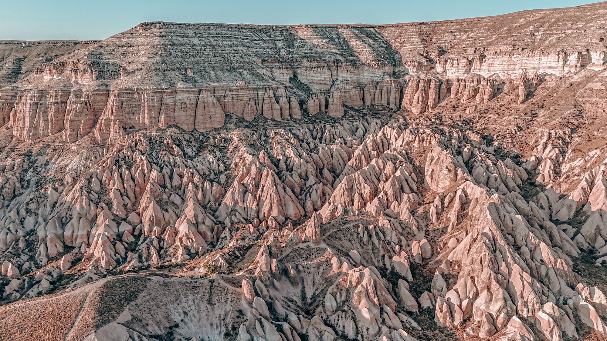 Aerial view of Cappadocia’s Rose Valley with jagged pink rock spires, steep ridges, and a towering cliff face under a clear blue sky