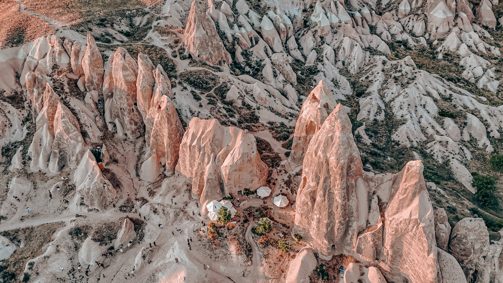 Aerial view of the dramatic rock formations in Red and Rose Valley, Cappadocia, Turkey, with tall jagged cliffs in pink and beige hues rising from the valley floor, small white parasols marking a refreshment stop nestled between the rocks, and winding trails scattered with hikers exploring the landscape