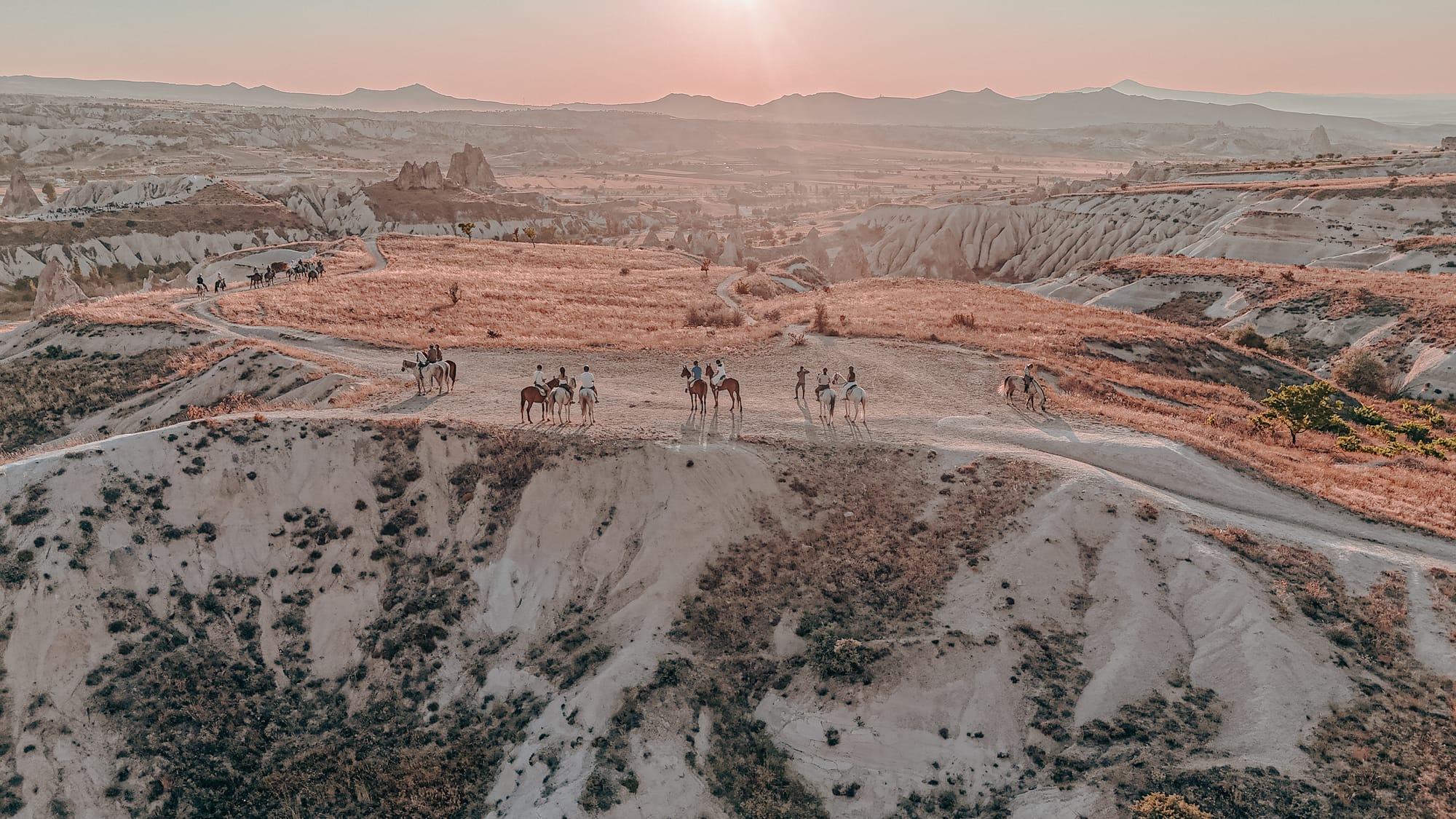 A group of horseback riders traveling along a winding trail at sunset in Cappadocia’s Red and Rose Valleys, surrounded by soft pink and beige rock formations and rolling hills in the background