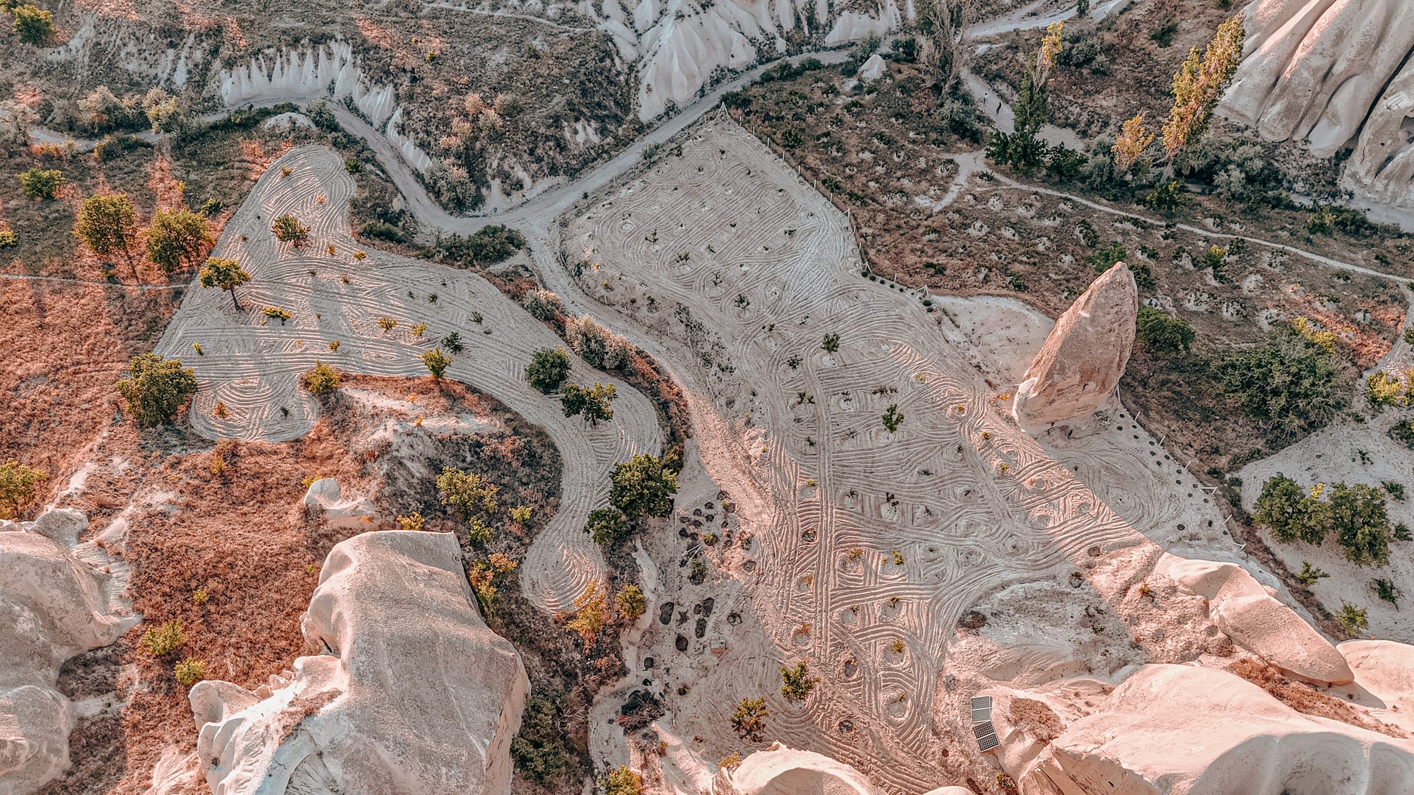 Aerial view of a valley in Cappadocia showing geometric plowing patterns in the sandy soil, surrounded by rock formations, scattered trees, and pathways winding through the landscape