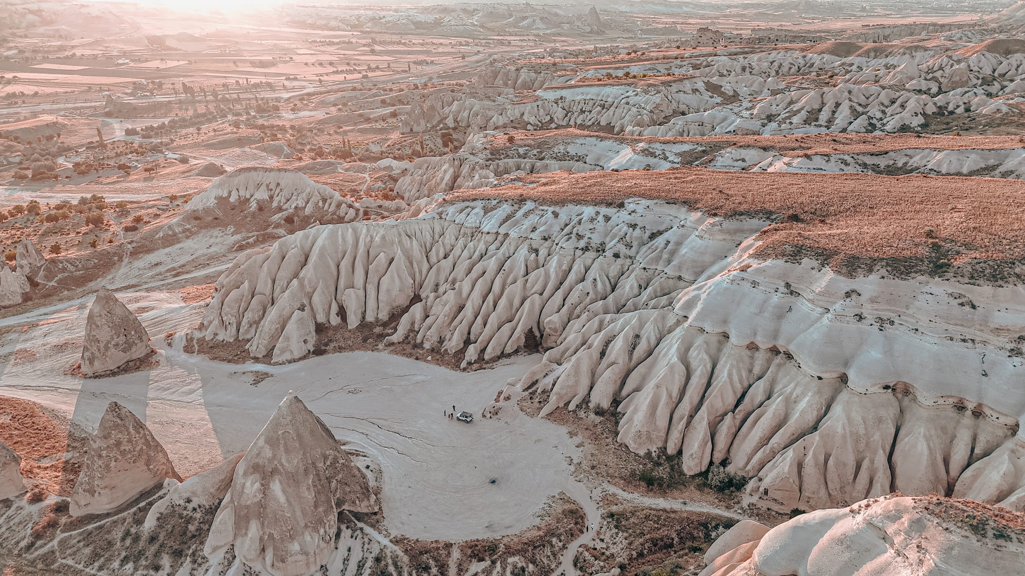 Aerial view of Cappadocia’s unique rock formations and ridged valley walls under the soft glow of morning light, with a few people and a vehicle visible in a flat sandy clearing surrounded by towering stone spires