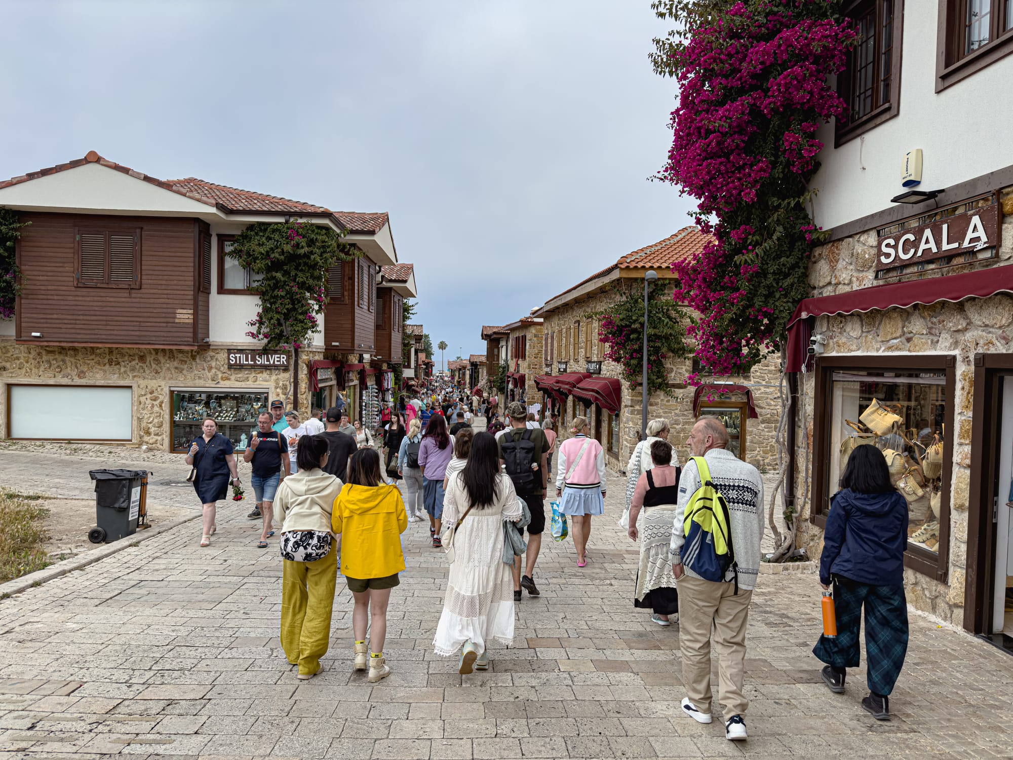 Crowds walking through the stone-paved main street of Side, Turkey, with shops on either side and blooming bougainvillea trailing from balconies above