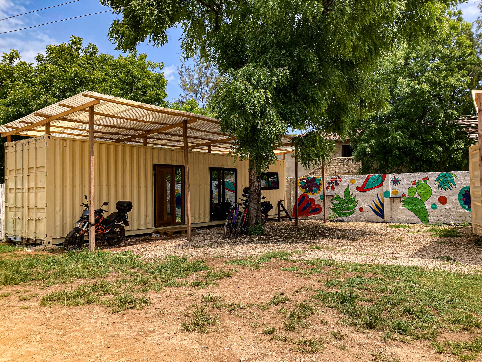 A cream-colored shipping container converted into a coworking space at Skippers Coliving in Diani Beach, with a corrugated roof extension, motorbikes and bicycles parked outside, and a colorful mural of plants and abstract shapes painted on the boundary wall under the shade of tall trees