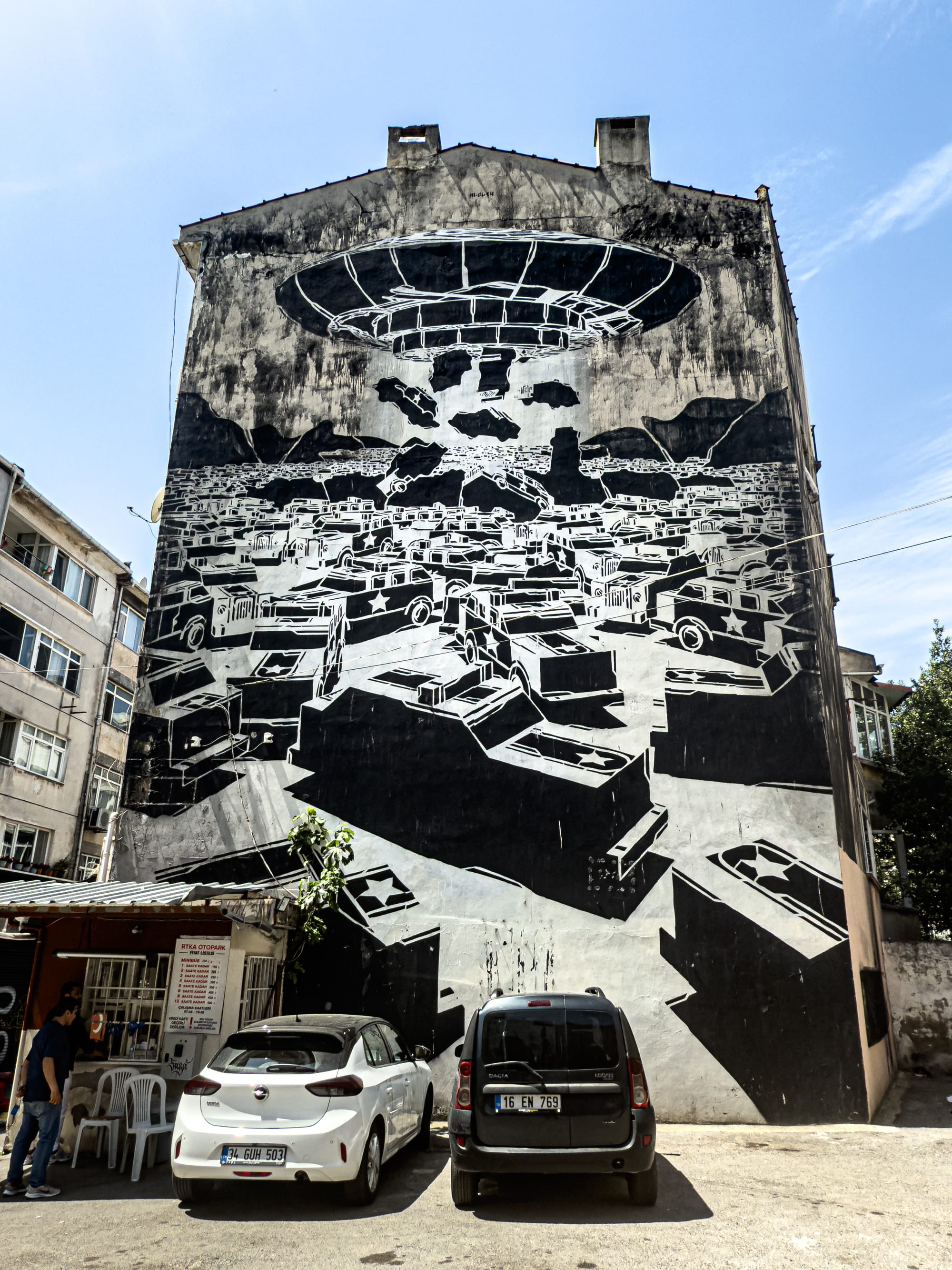 Black-and-white mural by M-City in Kadıköy showing a stylized aerial view of boxy military vehicles or tanks under a massive hovering structure, blending dystopian architecture with sci-fi symbolism