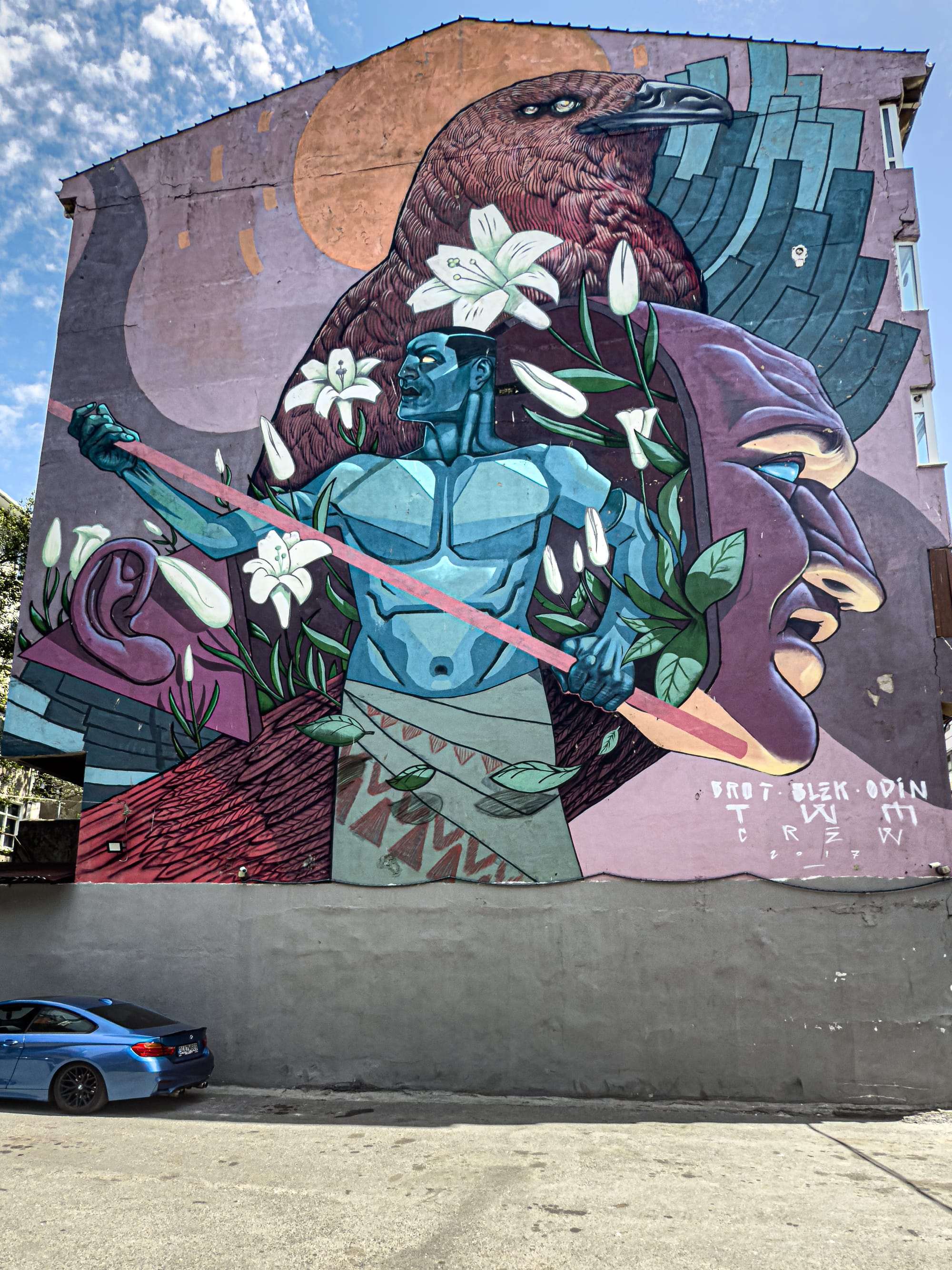 Large mural featuring a stylized blue male figure holding a spear, surrounded by lilies, disembodied ears and faces, and a giant bird with extended wings, set against a purplish backdrop on the side of a building