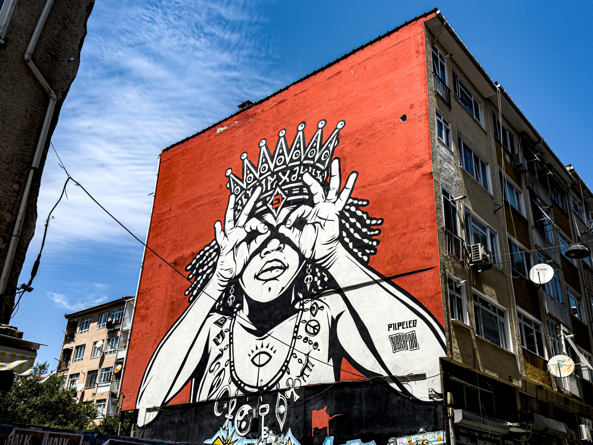 Large mural by Pilpeled showing a stylized Black woman wearing a crown and jewelry, holding her fingers in a circle over her eyes in a binocular gesture, painted in black and white on a bright red wall
