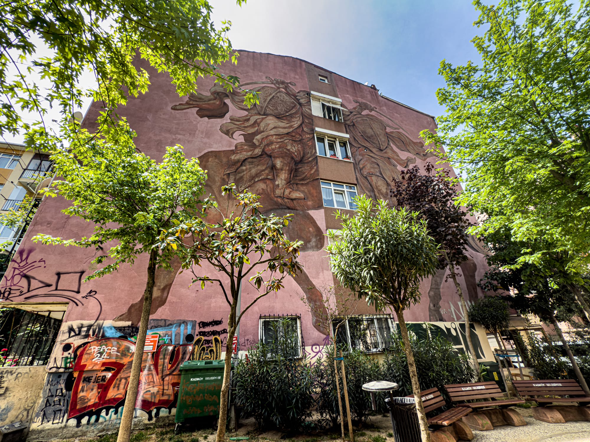 Large mural by Franco Fasoli on a pink building depicts two armored figures on horseback in dynamic motion, their forms mirrored and partially obscured by windows and balconies, framed by surrounding trees and urban graffiti
