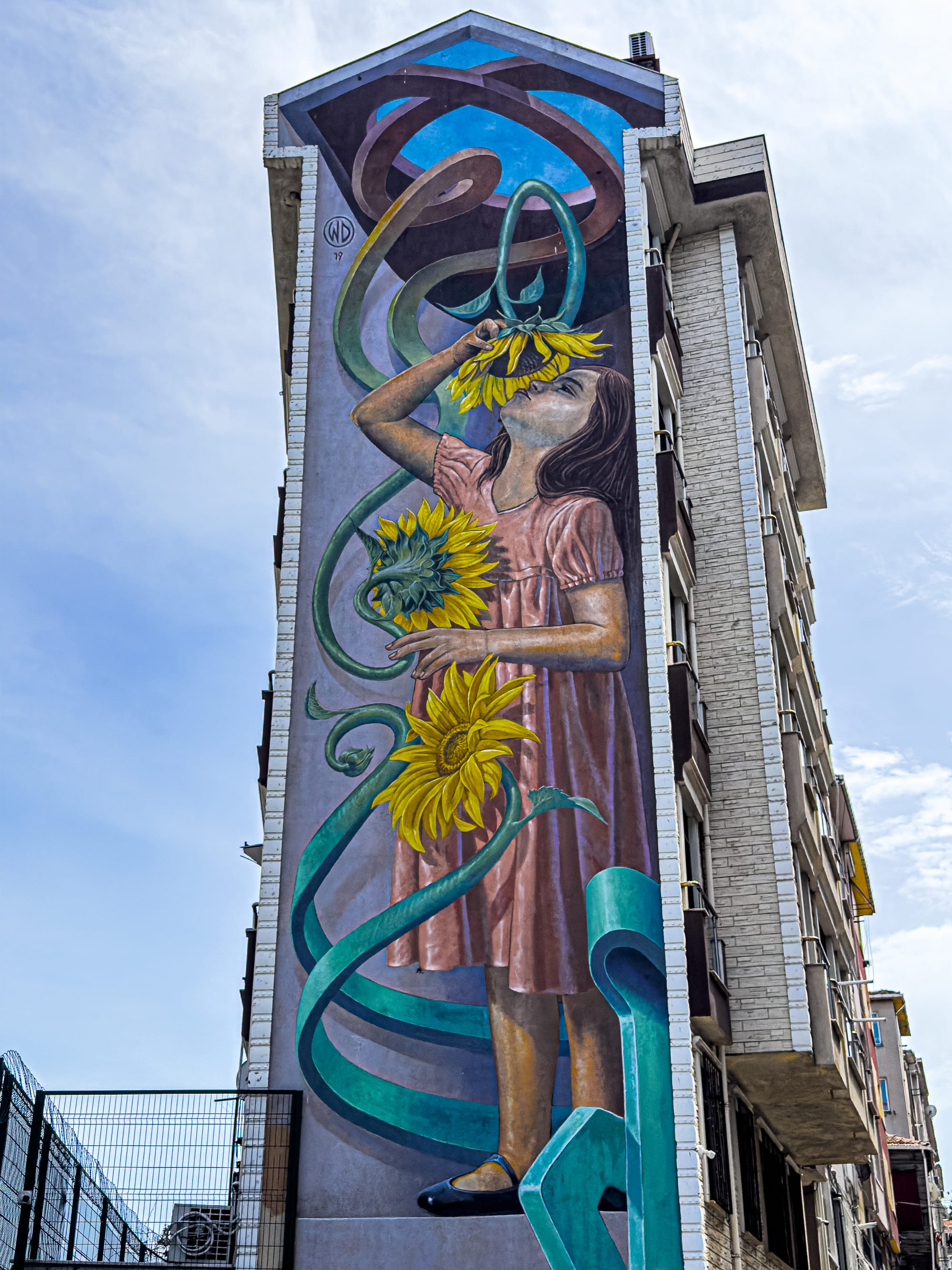 A tall mural by Indonesian artist WD depicts a girl in a pink dress holding and smelling vibrant yellow sunflowers, with swirling green stems and a backdrop of geometric sky tones, painted along the side of a high-rise in Kadıköy