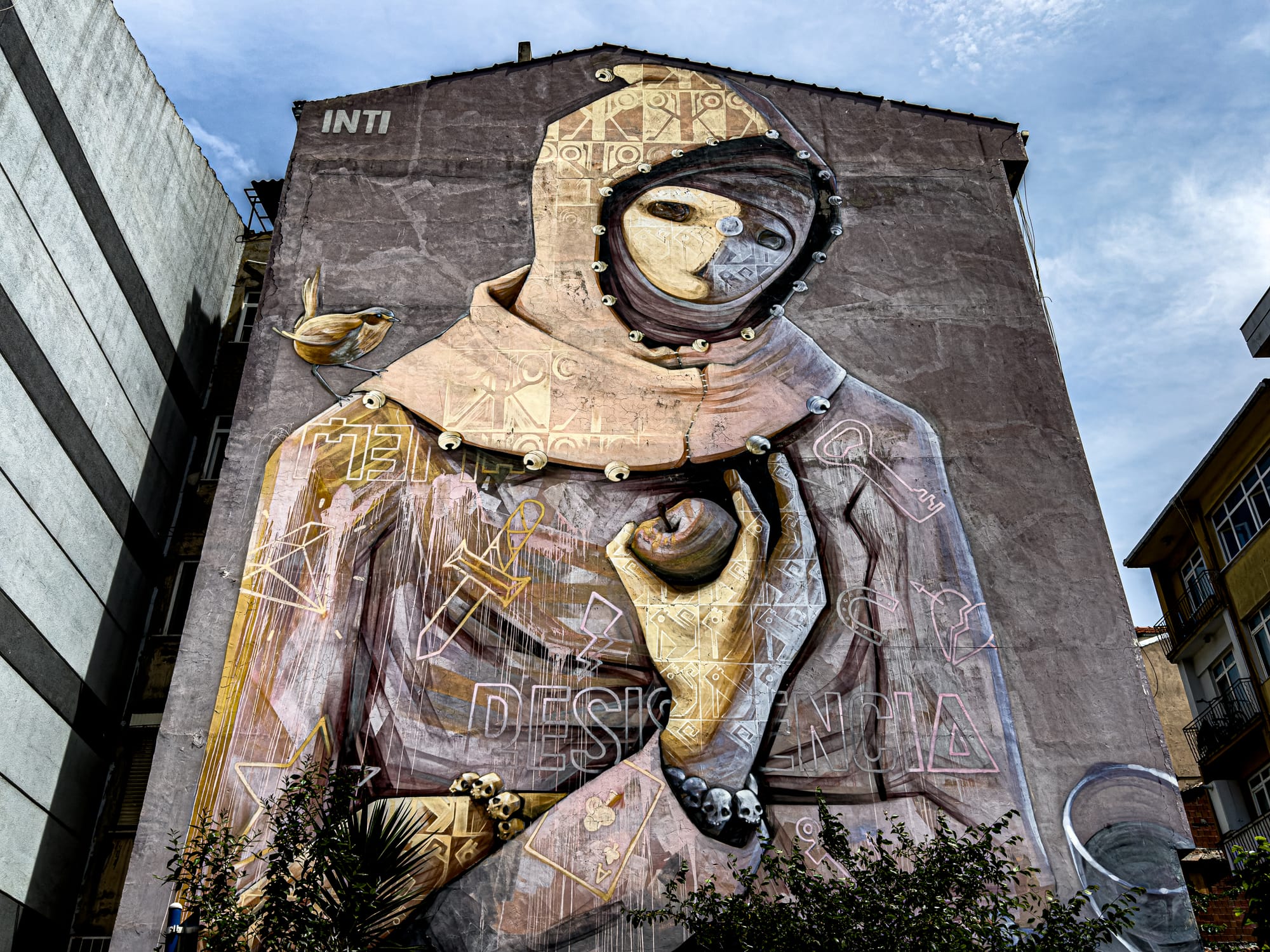 A monumental mural by Chilean artist INTI depicts a hooded, mask-like figure holding an apple, with symbols and skulls integrated into the cloak, painted on a multi-story building in Kadıköy