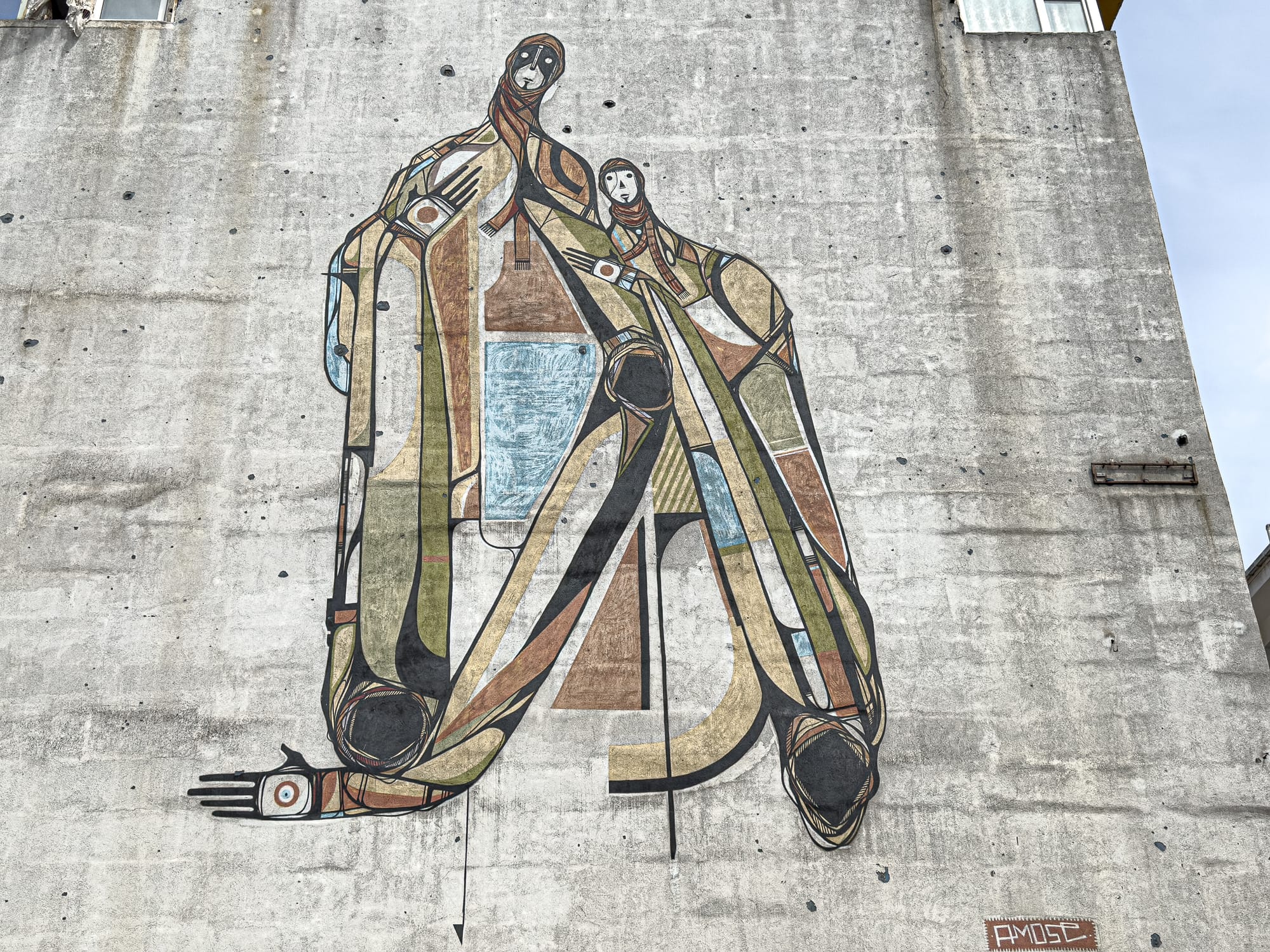 A large mural on a raw concrete wall features two abstract, elongated human figures intertwined, rendered in earthy tones and geometric patterns, with stylized faces and limbs stretched in fluid curves
