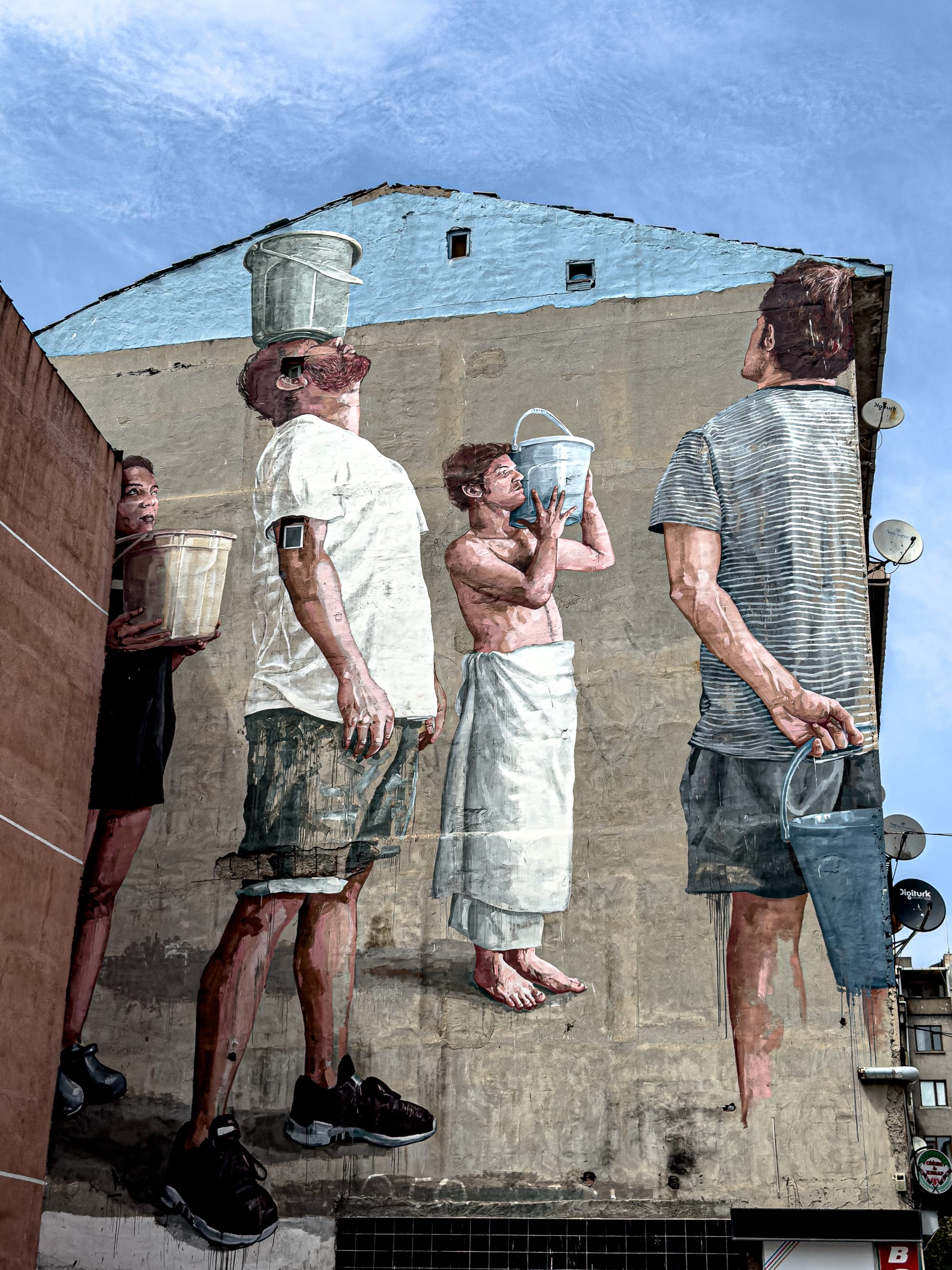 A large mural by Fintan Magee shows several figures holding buckets, one balancing it on his head and another drinking from one, painted in a realistic style on a tall building in Kadıköy