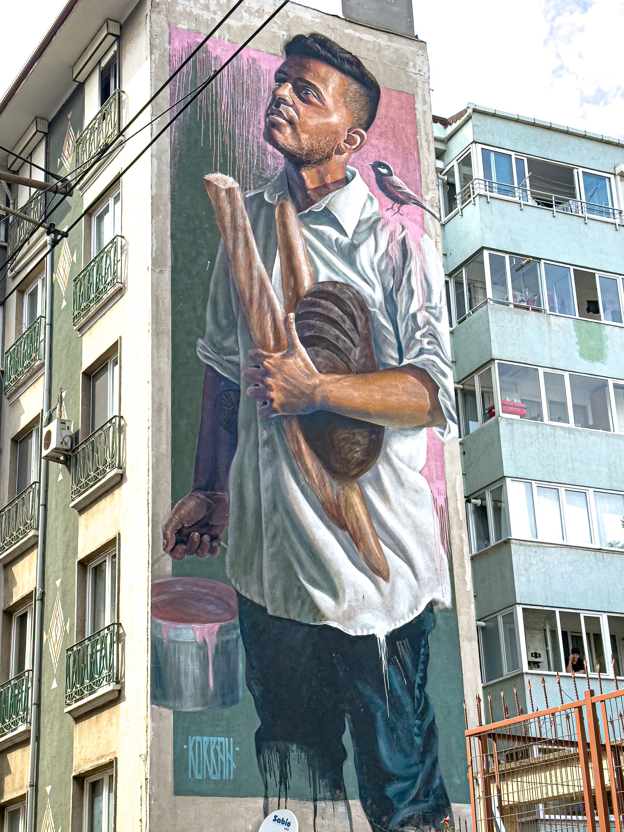 A large vertical mural in Kadıköy shows a man in a white shirt holding a baguette and a round loaf of bread, with a small bird perched on his shoulder and a soft pink and green background behind him