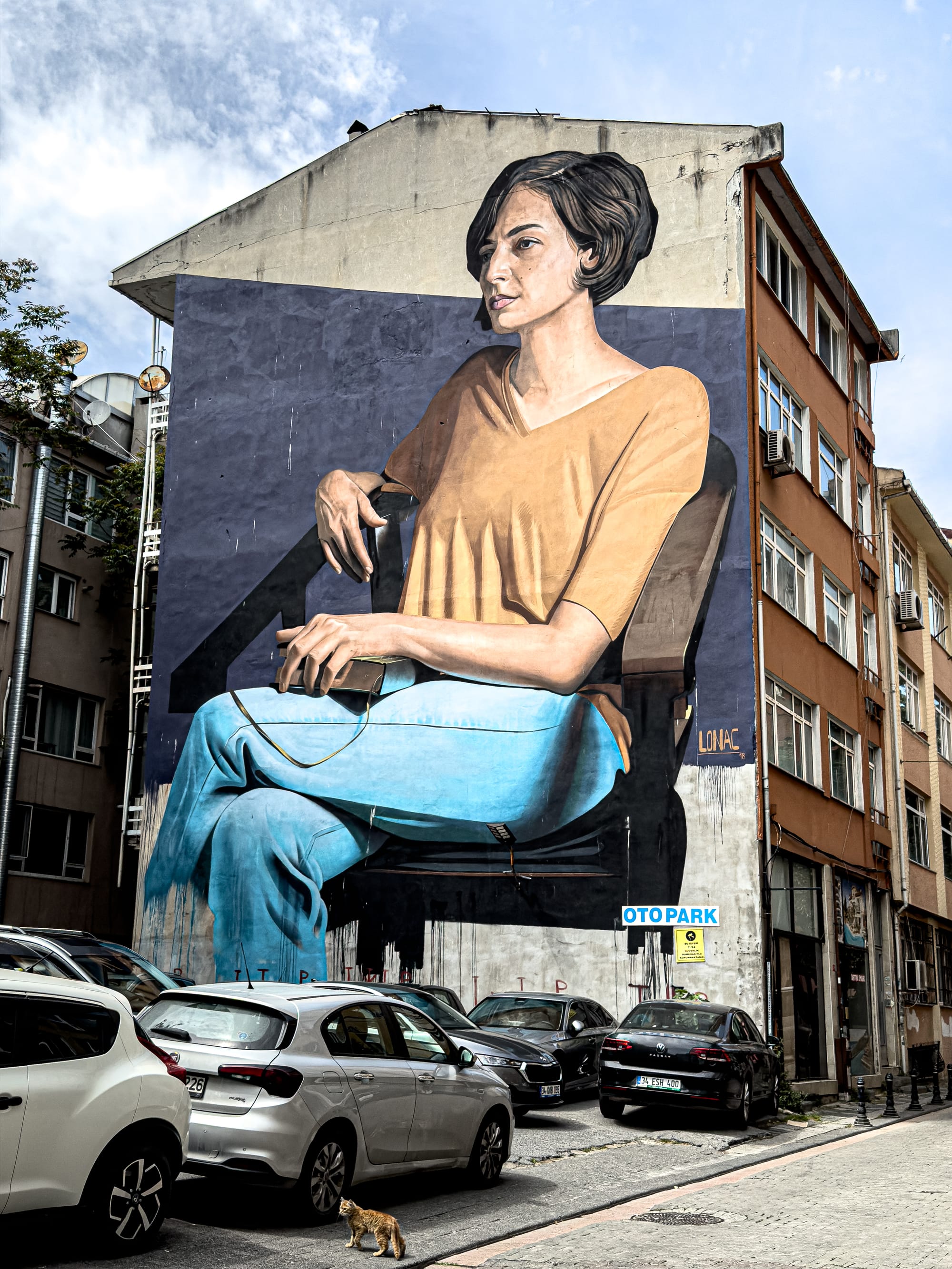 A large-scale mural depicts a woman with short dark hair, wearing a mustard shirt and blue jeans, sitting in a chair with one hand resting on her lap and the other holding a phone—painted with high realism against a dark blue background
