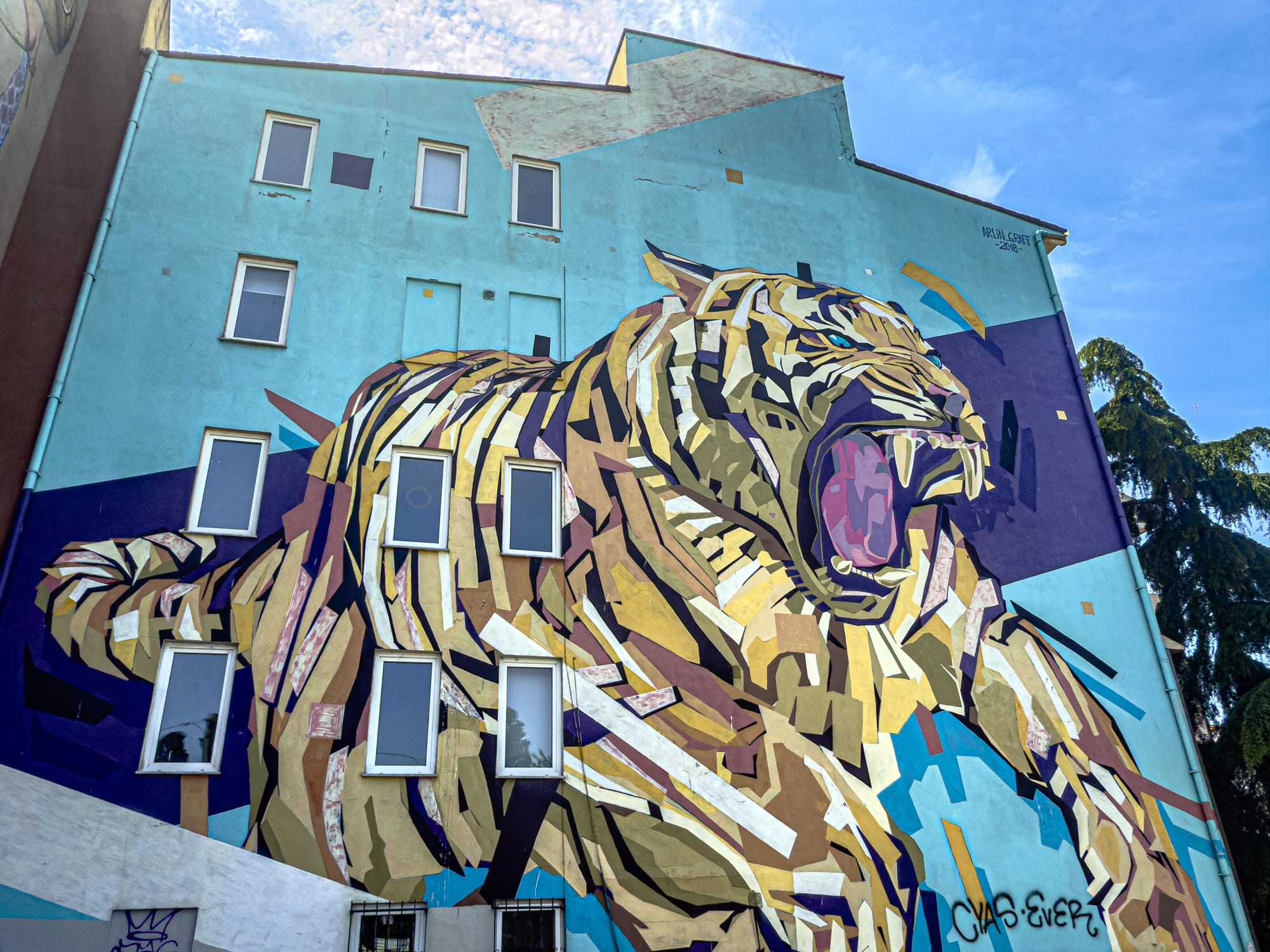 Street art mural of a stylized roaring tiger composed of sharp, angular geometric shapes in gold, cream, and purple tones, set against a teal-blue building wall with windows integrated into the design