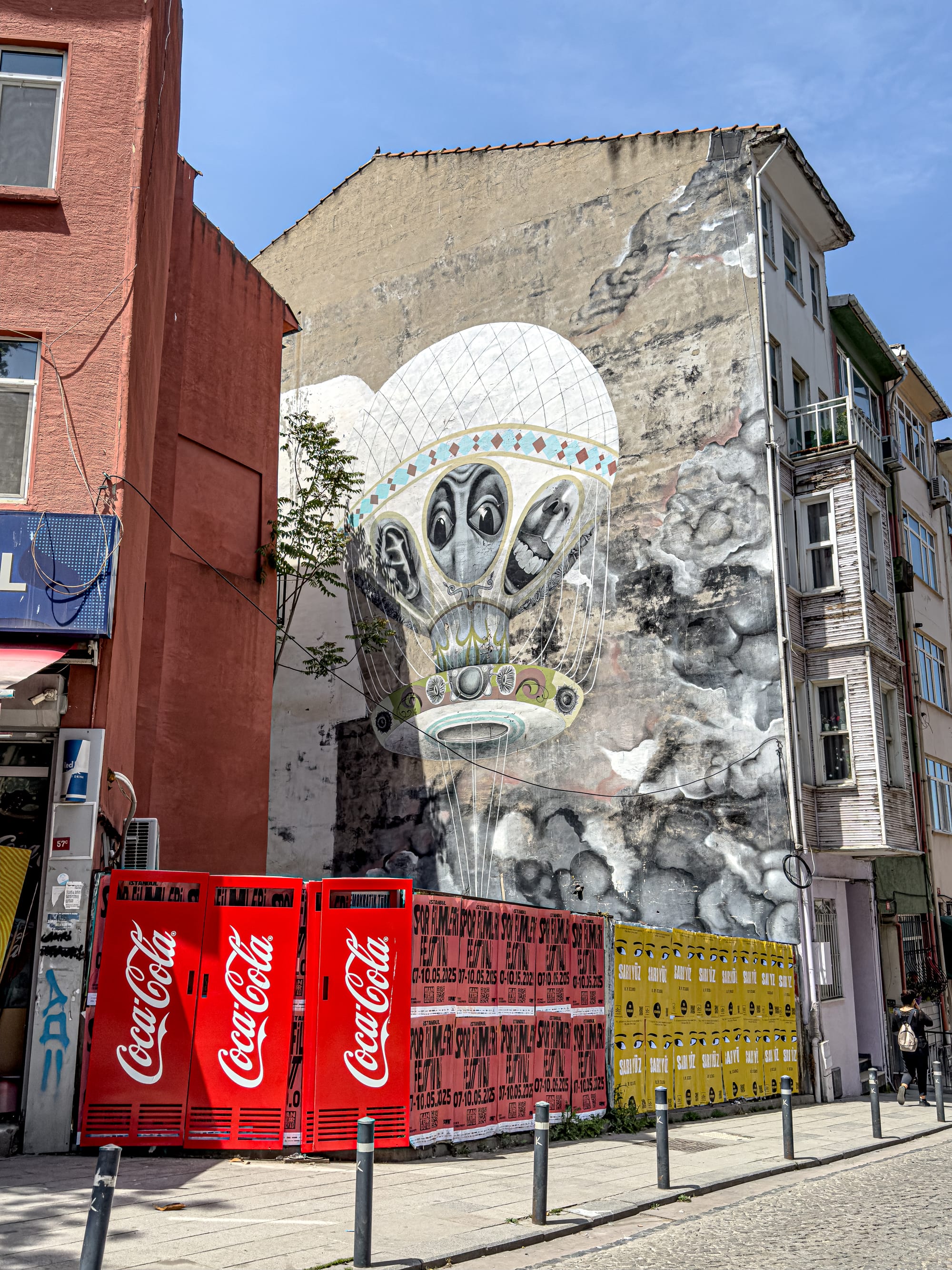 Mural of a large, monochromatic hot air balloon painted on a building; the balloon is tethered to a bed and features warped human faces on its basket, surrounded by cloudy, dreamlike textures