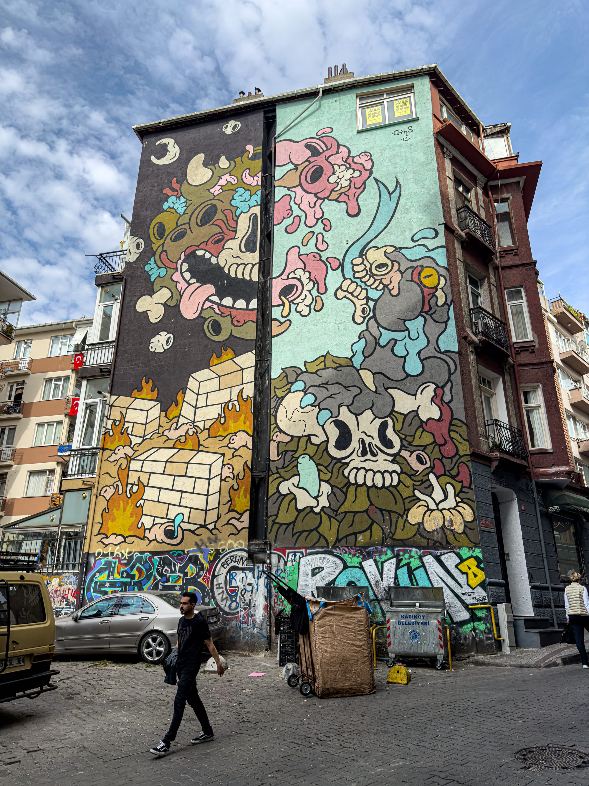 Tall, colorful street art mural by Turkish artist Cins featuring cartoonish skulls, melting brains, flames, cubes, and expressive characters divided across a black and mint-green background on the side of a multi-story building