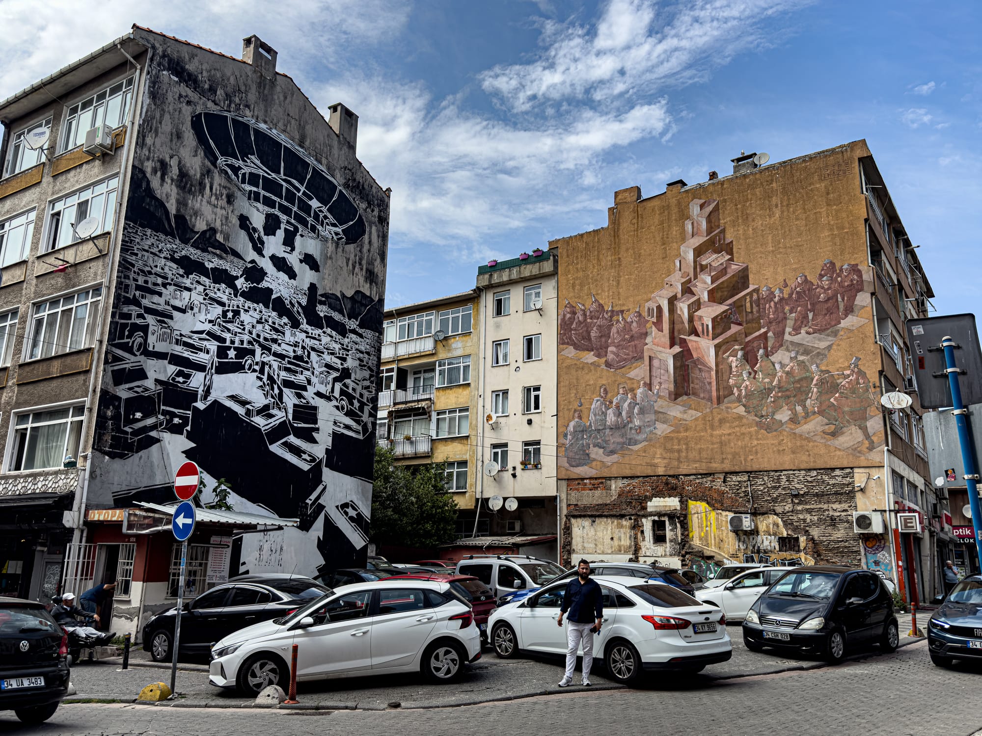A city corner in Kadıköy featuring two large-scale murals on adjacent buildings—one in black and white by M-City showing a grid of military vehicles under a hovering structure, and the other in earth tones by Sepe & Chazme depicting abstracted groups of people ascending toward a central architectural mass