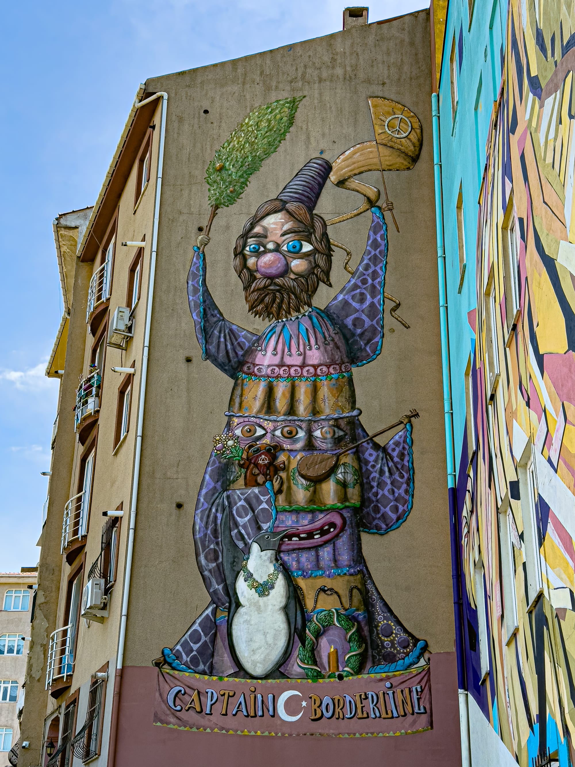 Tall mural by Captain Borderline showing a layered, fantastical totem figure with a bearded man in a wizard hat holding a tree and peace flag, stacked over multiple cartoonish faces, a teddy bear, a penguin wearing a flower necklace, and other symbolic elements