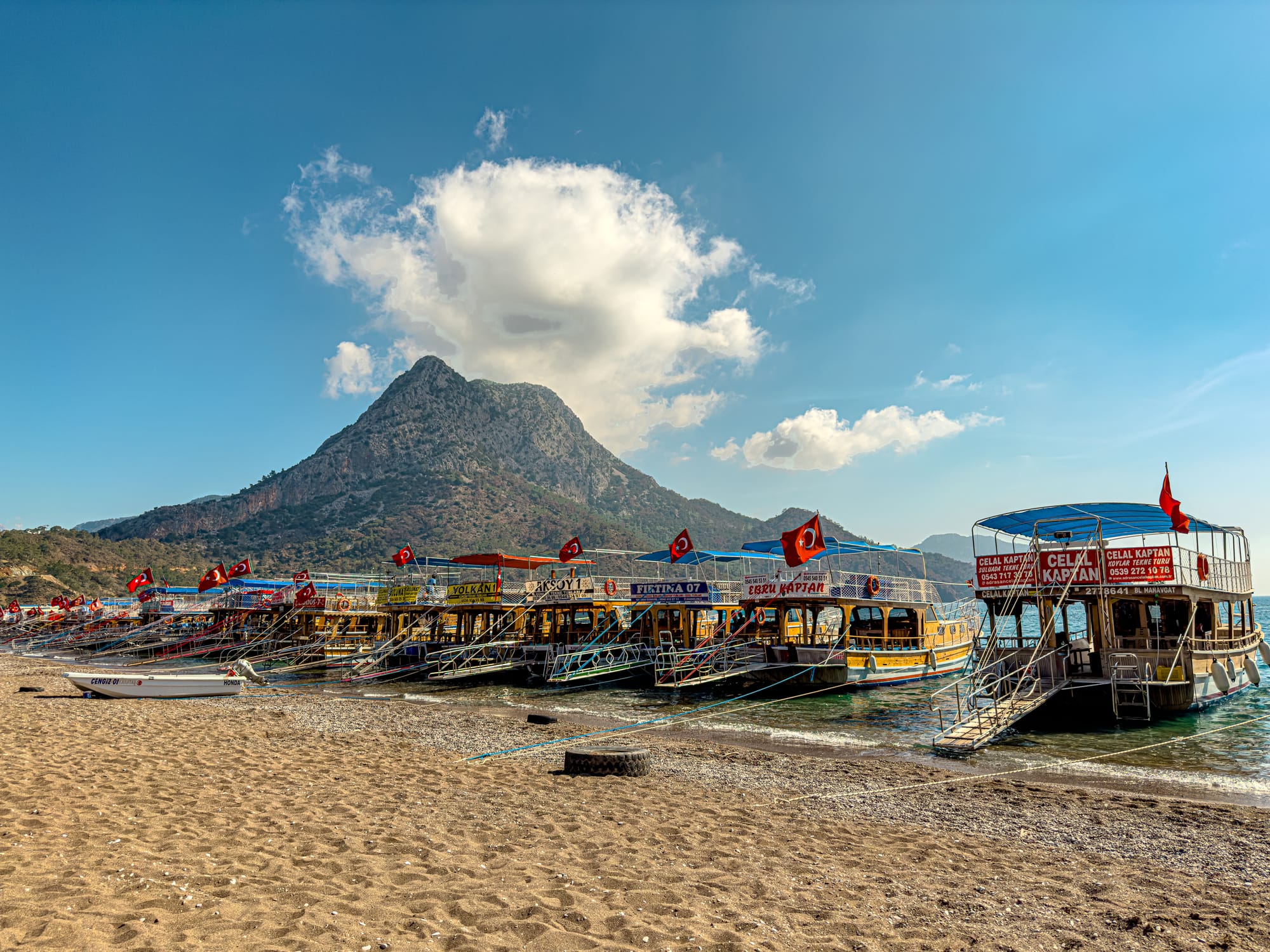 Colorful day-tour boats moored along the sandy beach in Adrasan, with a green-peaked mountain rising under a bright sky in the background