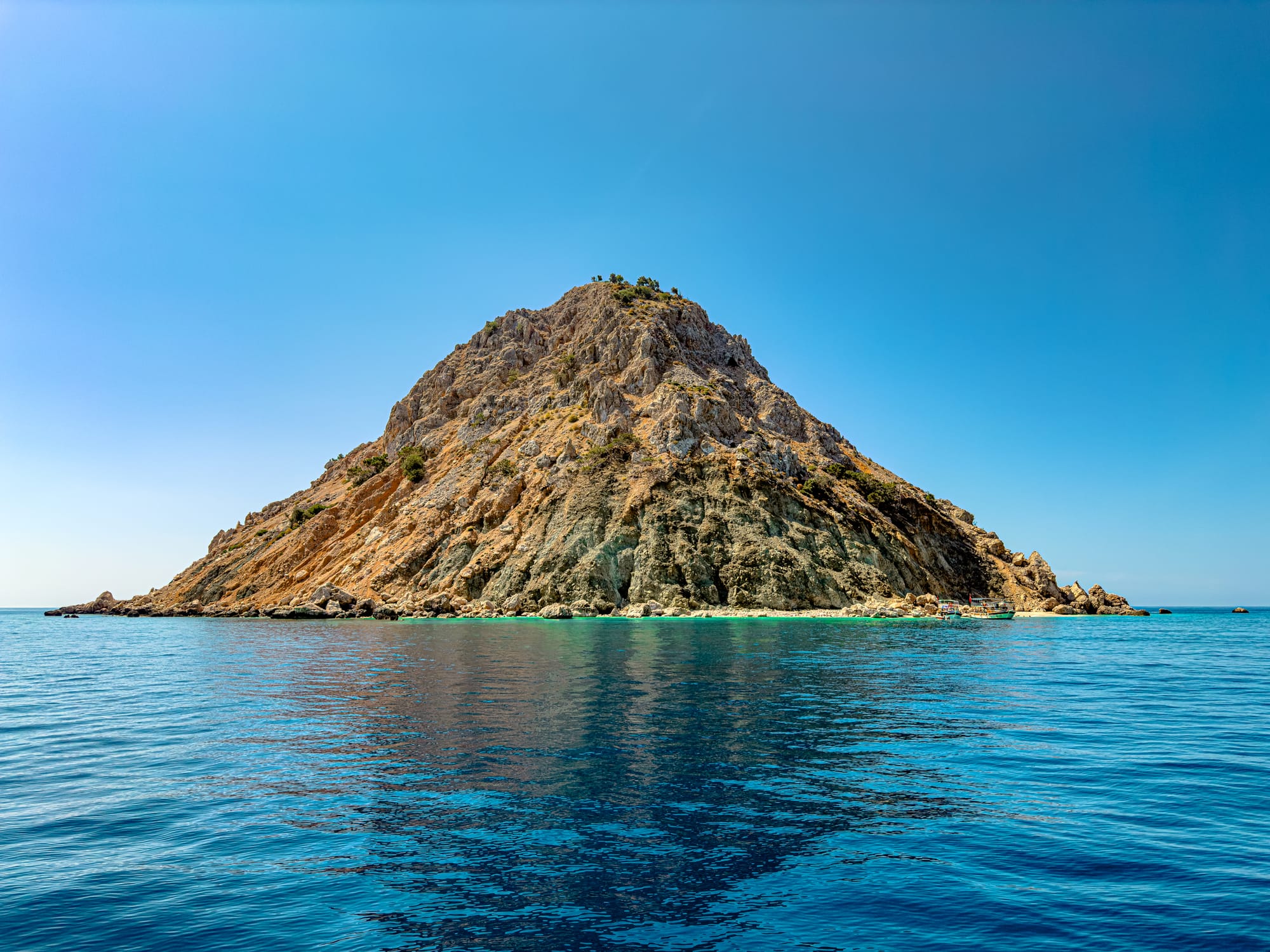 View of Suluada Island from the water, with its steep rocky slopes and turquoise shallows contrasting against the deep blue sea and clear sky
