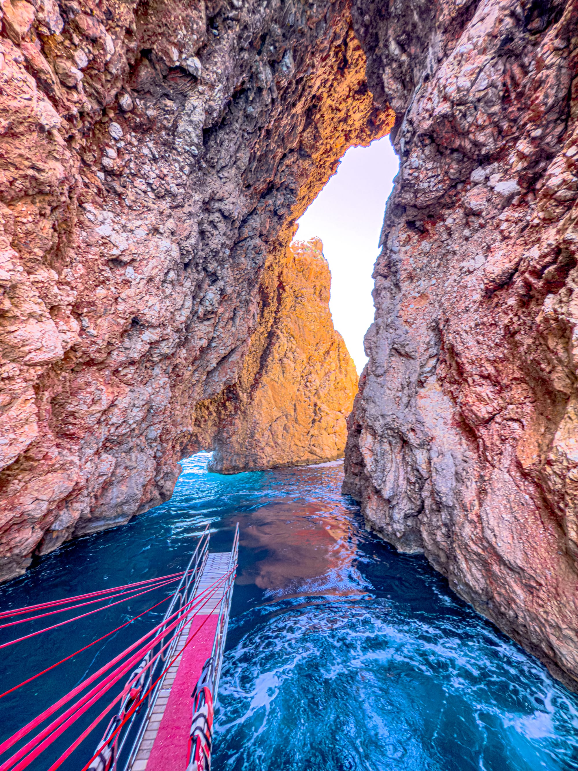 A boat passes through a dramatic, narrow sea gate between two towering rock cliffs near Suluada, with turquoise waves and warm light illuminating the passage