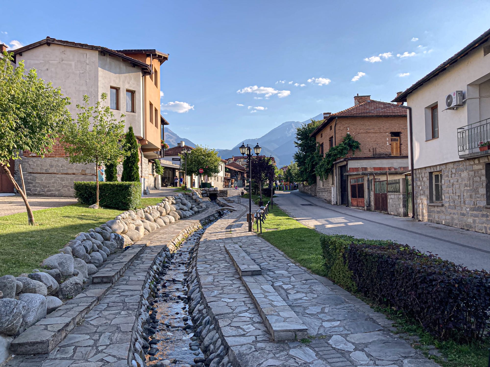 Narrow stone-lined water channel flowing through a quiet street in Bansko, with houses, greenery, and the Pirin Mountains in the background