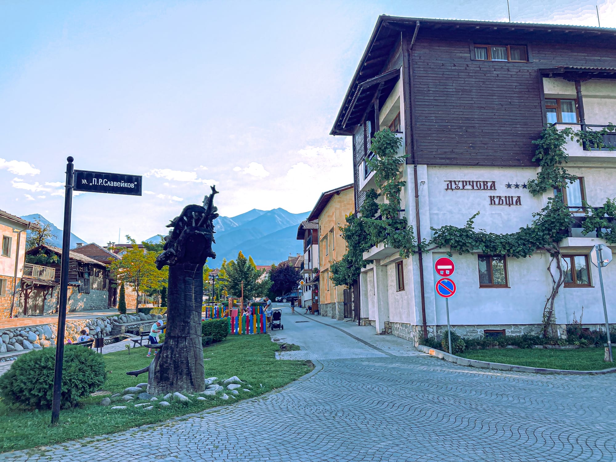 Quiet street in Bansko with a carved wooden sculpture, traditional houses covered in ivy, and mountains visible in the background