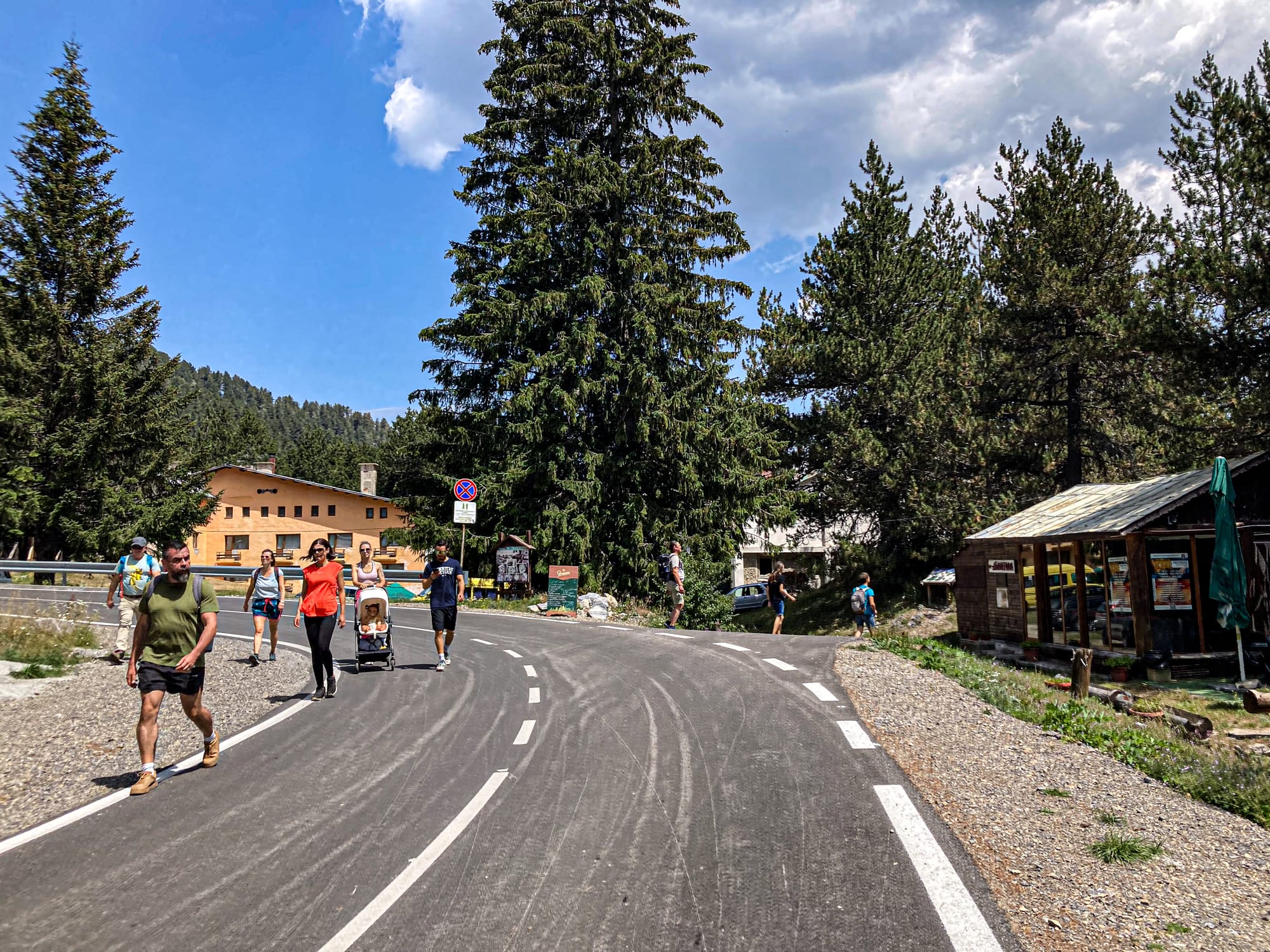 Groups of people, including a family with a stroller, walk along a paved road lined with tall pine trees near Vihren Hut in Bansko, Bulgaria, with a mountain lodge and small wooden structure visible in the background