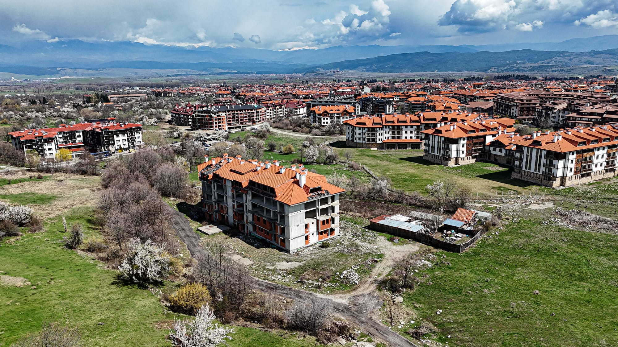 Aerial photo of Bansko, Bulgaria, showing a mix of completed apartment buildings with red-tiled roofs and abandoned or unfinished construction projects, with green fields and the Pirin and Rila mountains in the distance under a cloudy sky