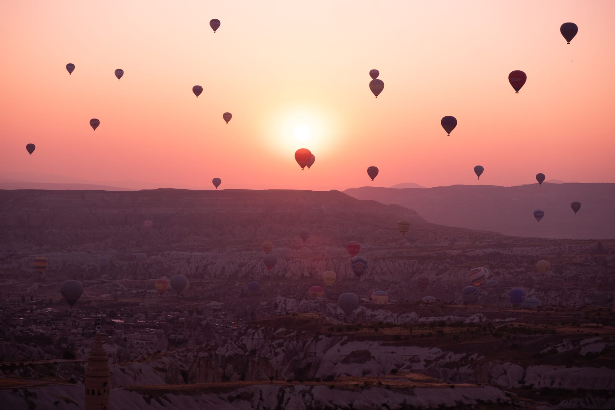 Sunrise view over Cappadocia, Turkey: the sun glows low on the horizon as dozens of hot air balloons float across the rocky valleys and ridges