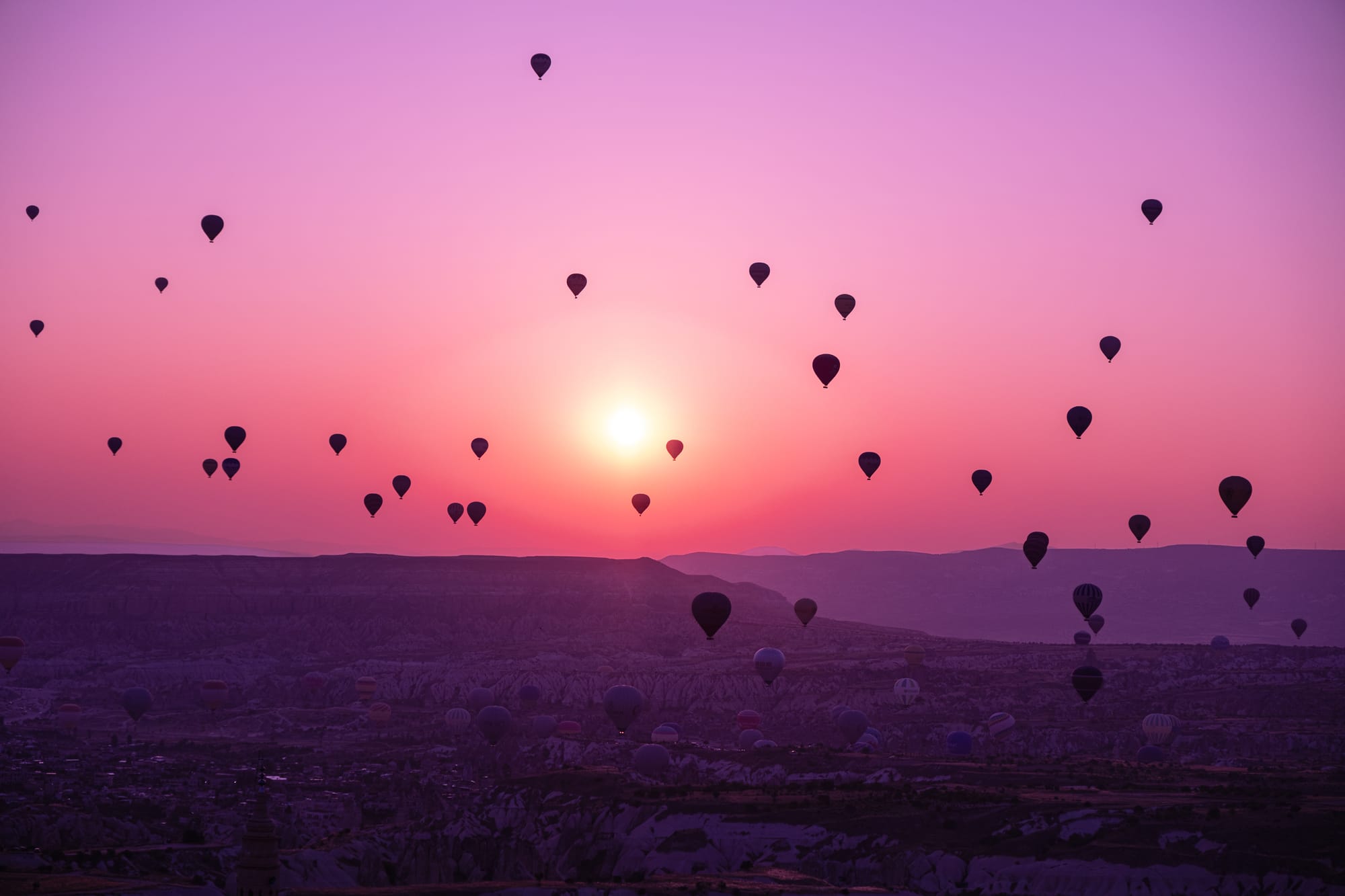 Sunrise over Cappadocia, Turkey, with dozens of hot air balloons floating above the ridged volcanic landscape