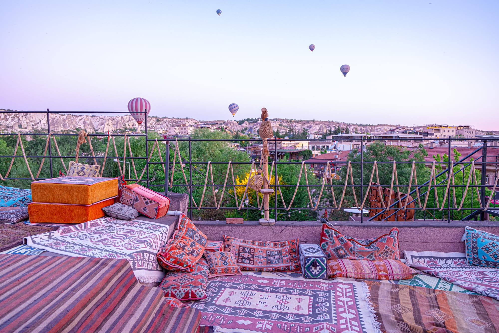 Colorful patterned rugs and cushions on a rooftop terrace in Cappadocia, Turkey, arranged for sunrise viewing. Beyond the railing, stone ridges and town rooftops stretch into the distance while several hot air balloons rise into the pale lavender sky