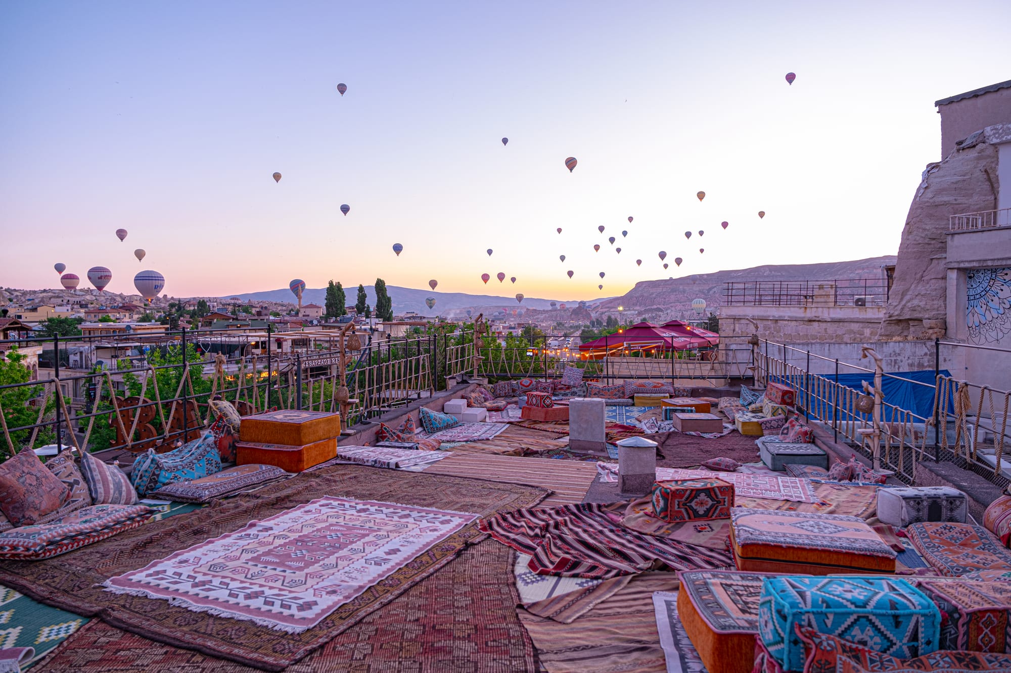 Sunrise from a rooftop in Göreme, Cappadocia: patterned rugs and floor cushions spread across the terrace, with dozens of hot air balloons drifting above the valleys and ridges in the background under a pale lavender sky