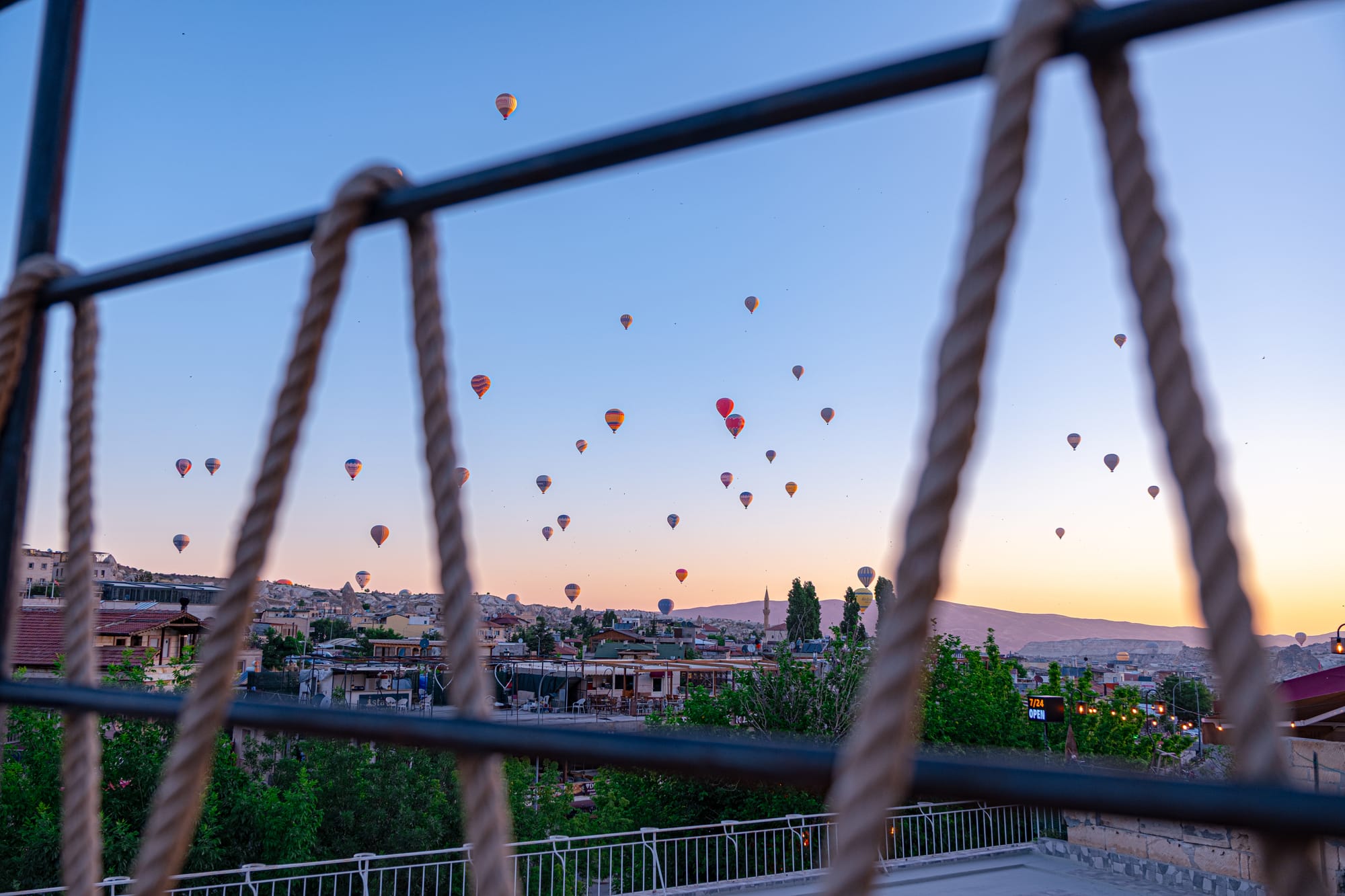 View of Göreme, Cappadocia, framed through the rope railing of a rooftop terrace. Beyond the ropes, dozens of colorful hot air balloons rise into the sky at sunrise, with stone houses, minarets, and ridges visible against a soft golden horizon